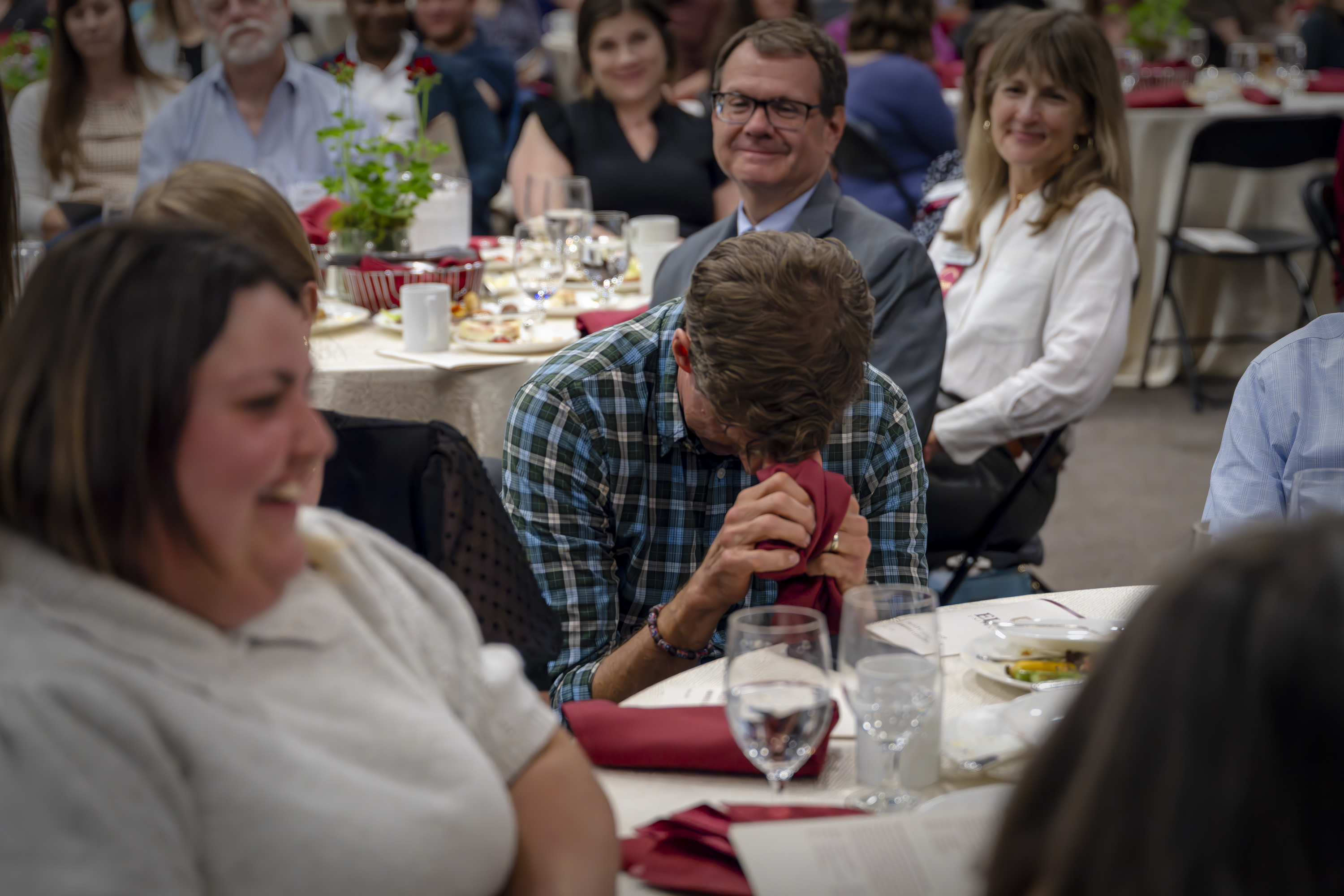 \A man sits at a banquet table visibly emotional, covering his face with a napkin, while others around him smile and look on supportively.