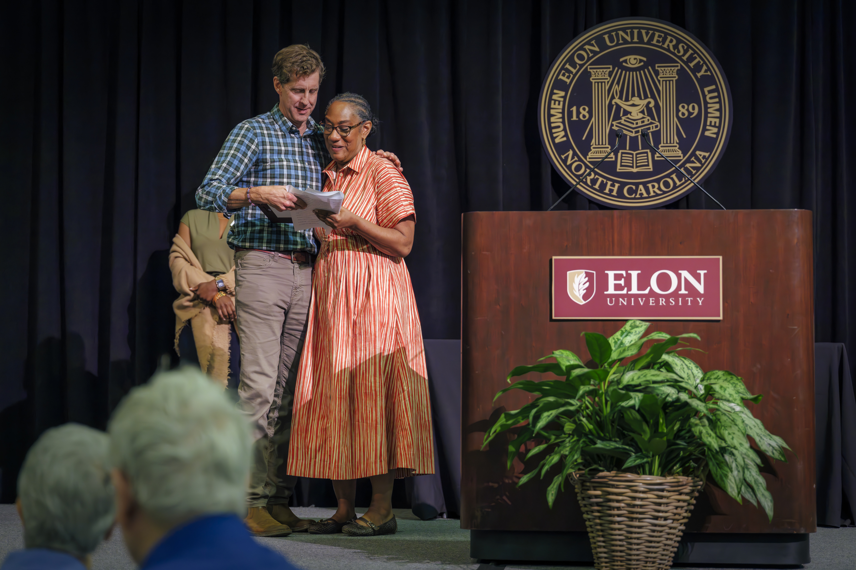 A man and woman stand together on stage at an Elon University event beside the podium with the university seal in the background.