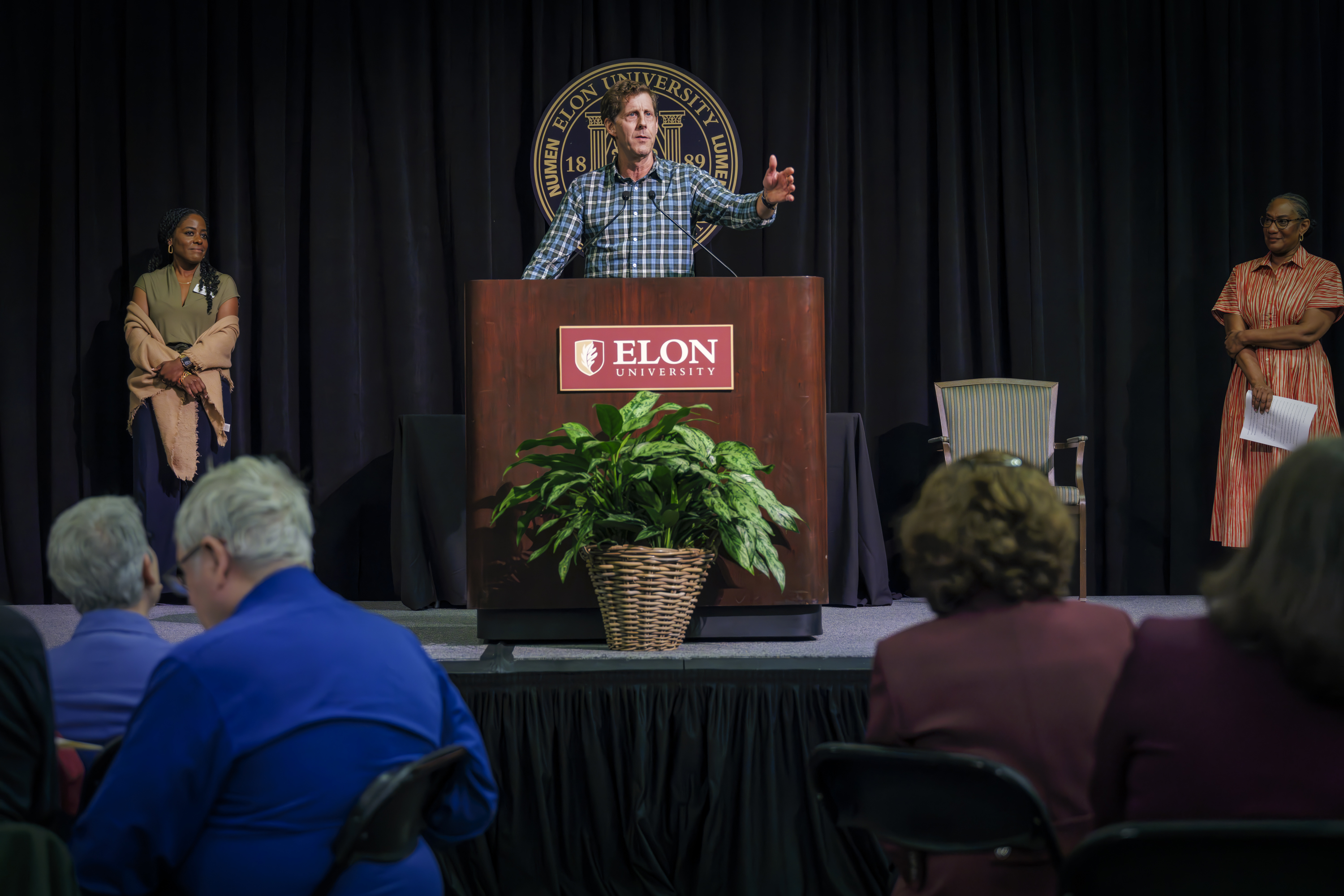 A man gestures expressively while speaking at the Elon University podium, as two women stand on either side of the stage listening attentively.