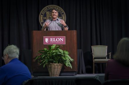 A man speaks at a podium with the Elon University logo and seal behind him, addressing an audience during a formal event.