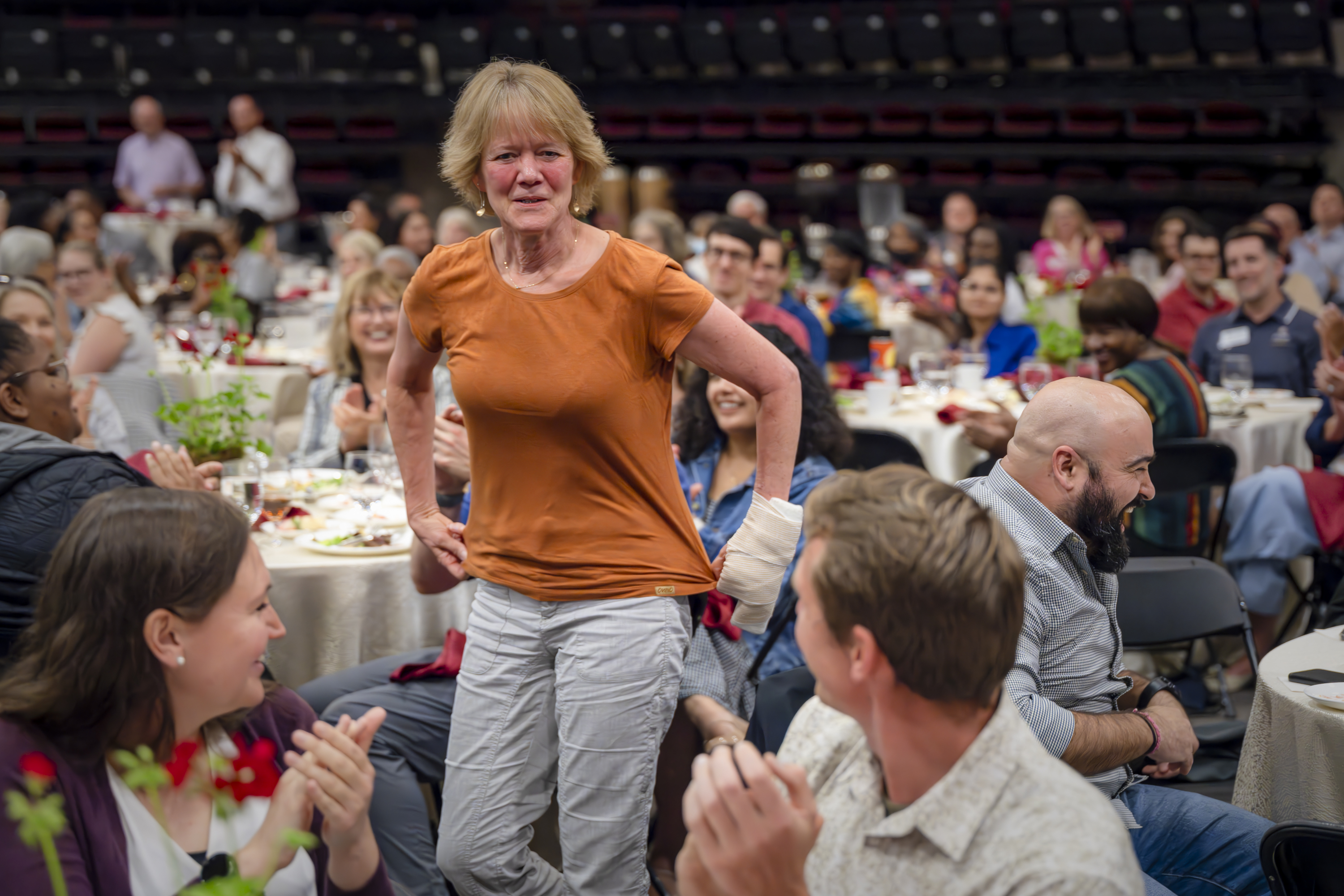 A woman stands smiling and emotional as people around her applaud during a formal event with round banquet tables and a large seated audience.