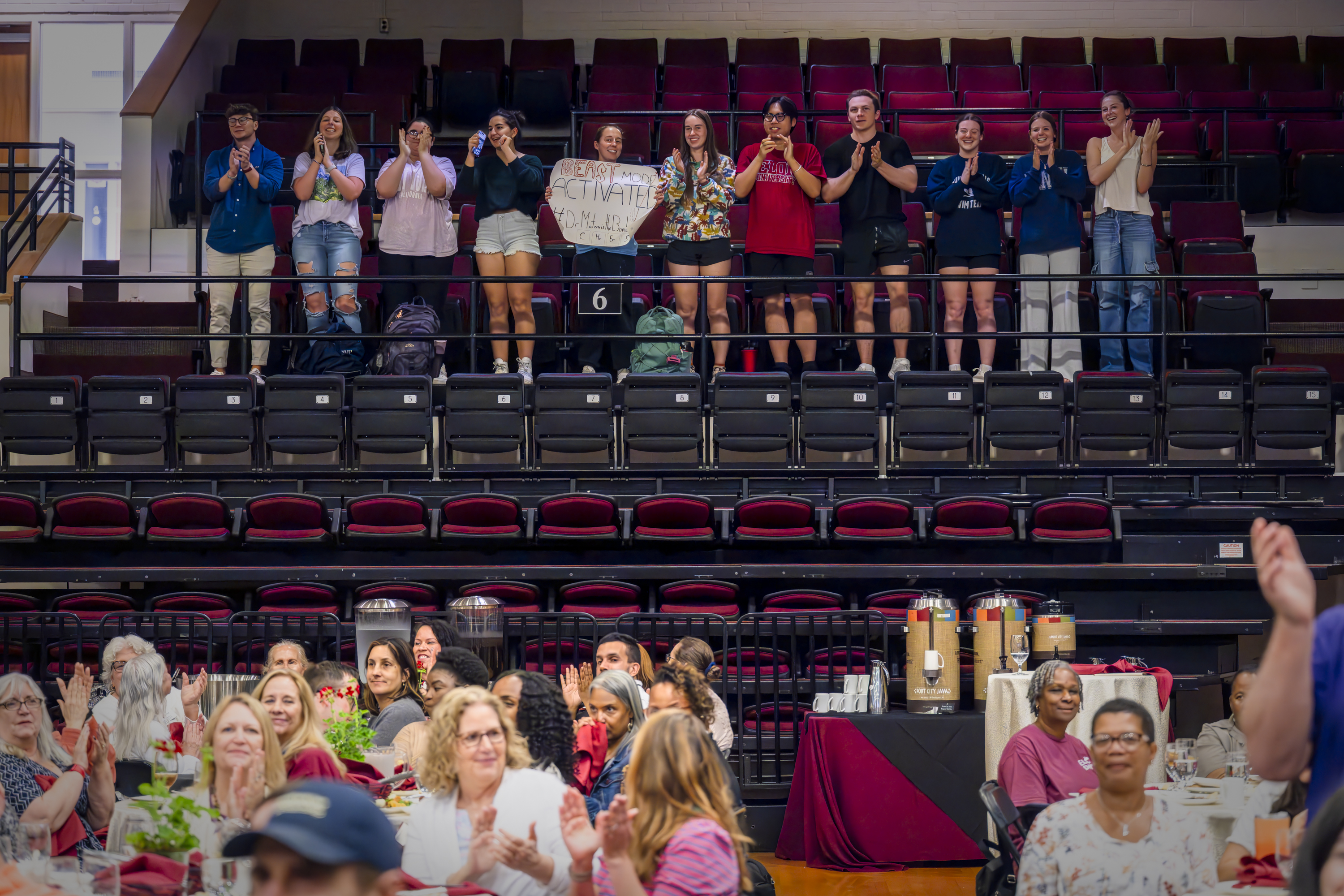 A group of students stands and cheers from the bleachers, one holding a handmade sign, while attendees at round tables below look on and applaud during an event.