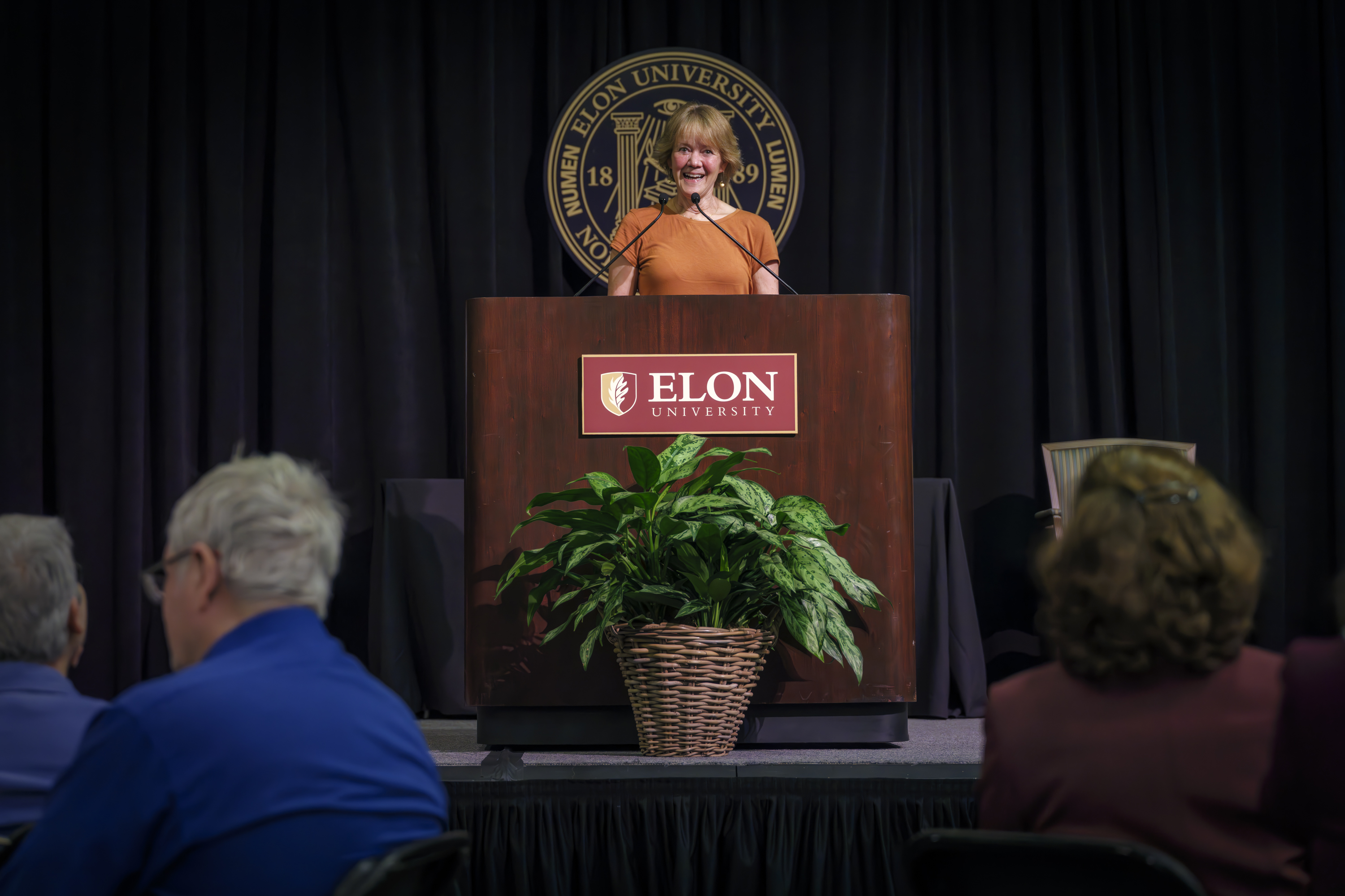 A woman stands at a podium with the Elon University seal behind her, speaking to an audience.