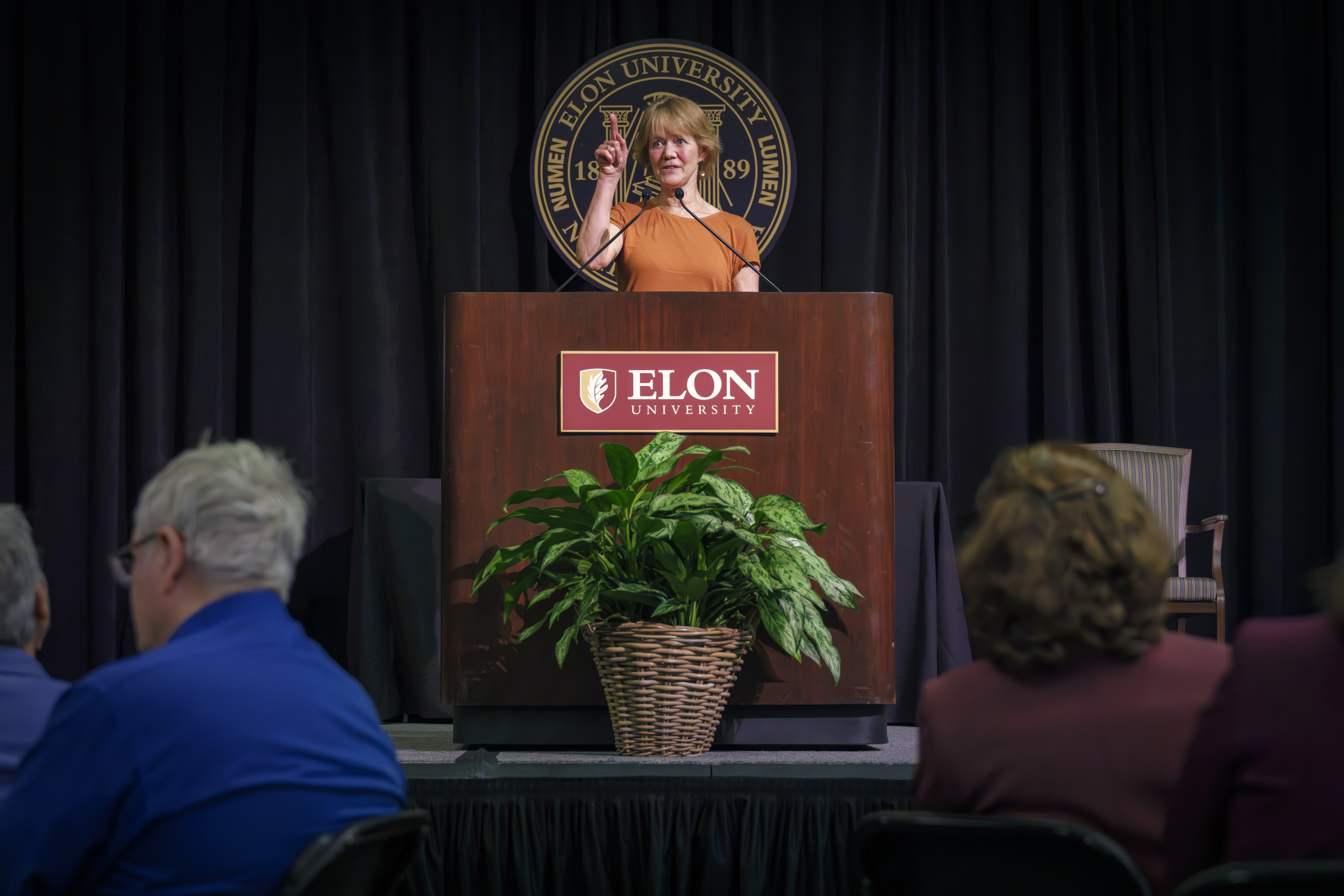 A woman stands at a podium with the Elon University seal behind her, speaking to an audience and gesturing with one hand raised.