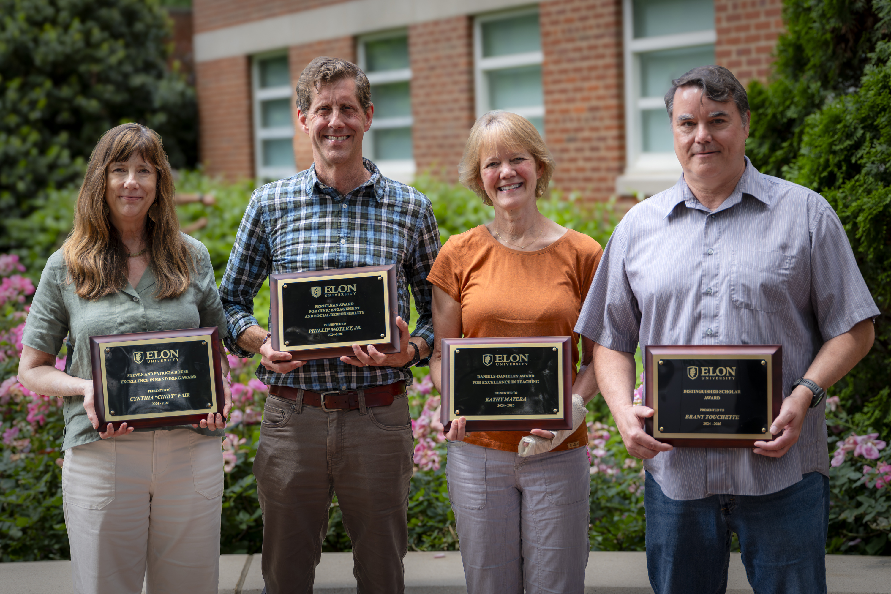 Four people stand smiling outside in front of a brick building and flowers, each holding a plaque from Elon University.