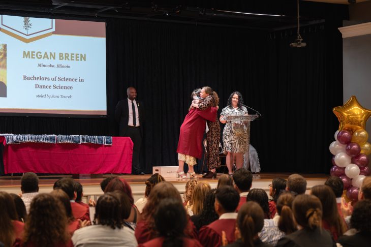 A graduate in a maroon gown embraces a presenter on stage during Elon University's First-Generation Stole Ceremony. The screen behind them displays the graduate’s name, Megan Breen, along with her hometown, Minooka, Illinois, and her degree: Bachelors of Science in Dance Science. The presenter is listed as Sara Tourek. Audience members watch and applaud while a table with blue stoles is visible beside them, and colorful balloons decorate the stage.