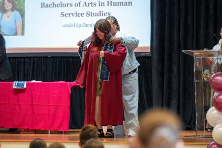 A graduate in a maroon cap and gown smiles as they are honored during the First-Generation Stole Ceremony. A presenter places a navy blue stole over their shoulders. Behind them, a large screen displays the graduate's name, degree in Human Service Studies, and the person bestowing the stole. The ceremony takes place on a stage decorated with maroon and white elements.