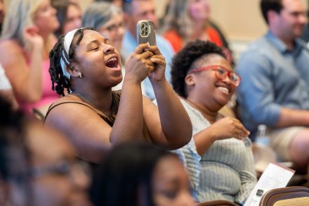 A cheering guest holds up a smartphone to record a special moment during the First-Generation Stole Ceremony. She is smiling widely and celebrating, surrounded by other joyful attendees seated in rows, some clapping and laughing.