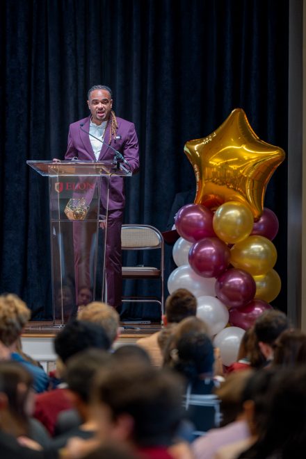A speaker stands at a clear podium with the Elon University logo, addressing an audience during the First-Generation Stole Ceremony. He wears a maroon suit and speaks confidently into the microphone. To the right of the podium, a decorative balloon arrangement in gold, maroon, and white forms a star-shaped display. Audience members are visible in the foreground, attentively listening.