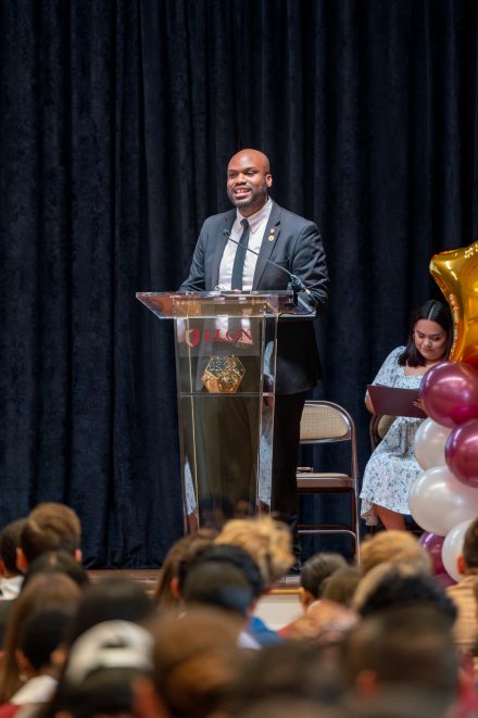A smiling speaker stands at a clear podium with the Elon University logo during the First-Generation Stole Ceremony. He is dressed in a dark suit and tie, and behind him sits a woman in a floral dress reviewing notes. A festive balloon arrangement in gold, maroon, and white is visible to the right, with a large audience seated in front.