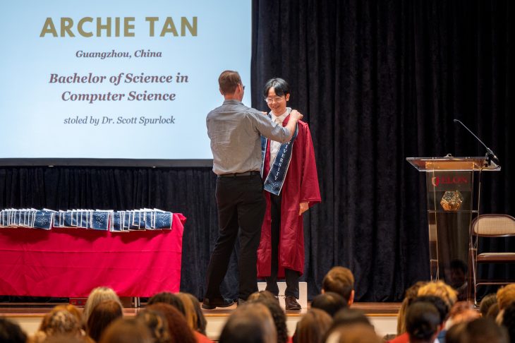 A student wearing a maroon graduation gown smiles as a faculty member places a stole around their shoulders during Elon University's First-Generation Stole Ceremony. The large screen behind them displays the graduate’s name, “Archie Tan,” along with their hometown, degree in Computer Science, and the name of the faculty member bestowing the stole, Dr. Scott Spurlock. A table nearby holds more stoles, and the stage is set for a formal recognition event.