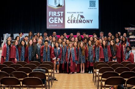 A large group of first-generation college graduates pose together on stage at Elon University’s First Gen Stole Ceremony. They are wearing maroon graduation gowns and navy stoles that read "ELON UNIVERSITY FIRST GEN." A screen in the background displays the event title and a QR code for graduate bios. Rows of empty chairs are visible in the foreground.