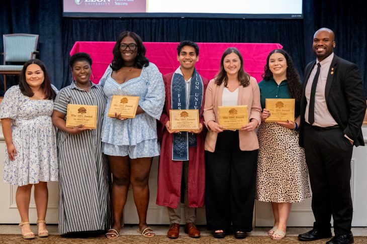 Seven individuals pose together on stage, smiling and holding wooden plaques that read "The 1891 Award." One recipient stands in the center wearing a maroon graduation gown and a navy stole with "FIRST GEN" and "ELON UNIVERSITY" embroidered on it. The group represents the honorees and organizers of the First-Generation Stole Ceremony, celebrating contributions to first-generation student success.