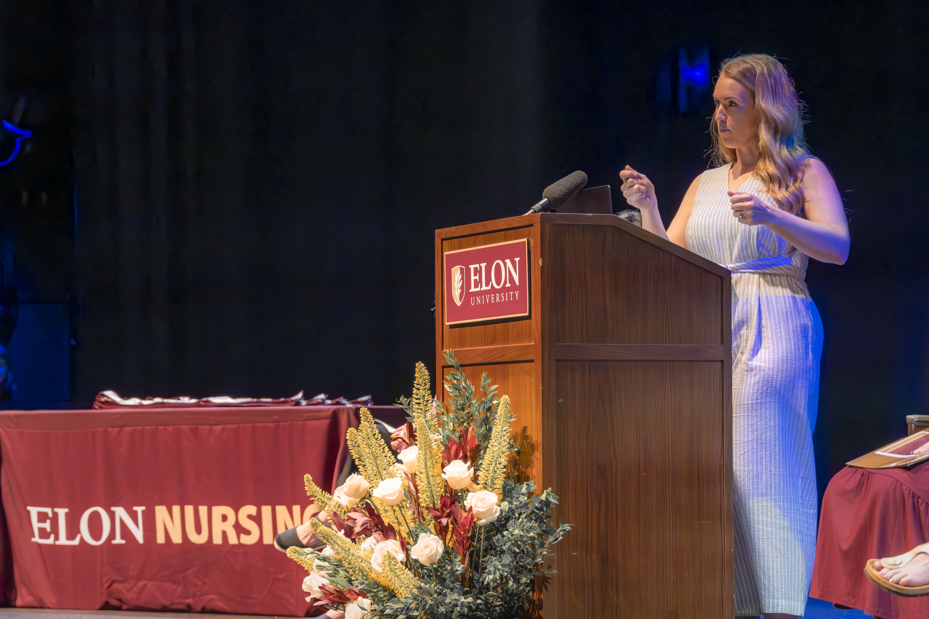 A speaker addresses the audience from a podium flanked by seated faculty and staff during an Elon Nursing event.