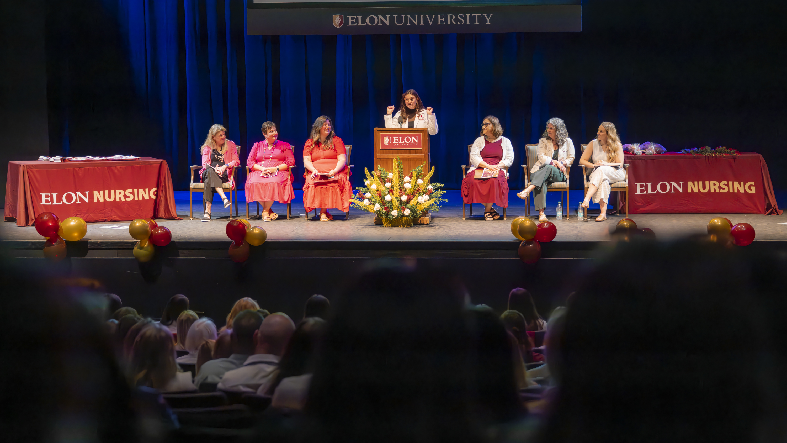 A woman stands at a podium labeled “Elon University” speaking during a ceremony on a stage decorated with flowers and an “Elon Nursing” tablecloth.