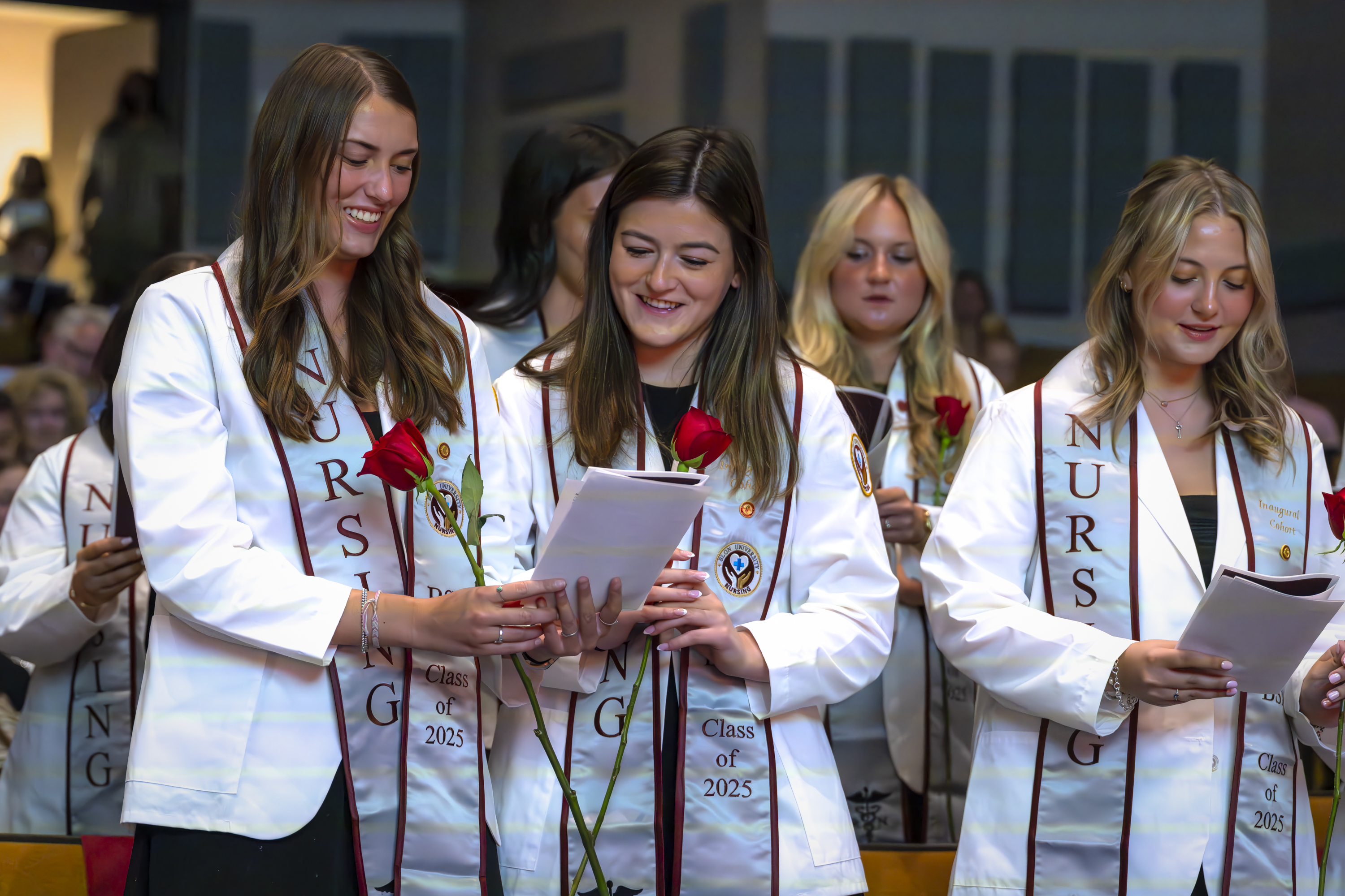 Nursing students in white coats and stoles labeled “Class of 2025” read from programs while holding red roses during a ceremony.