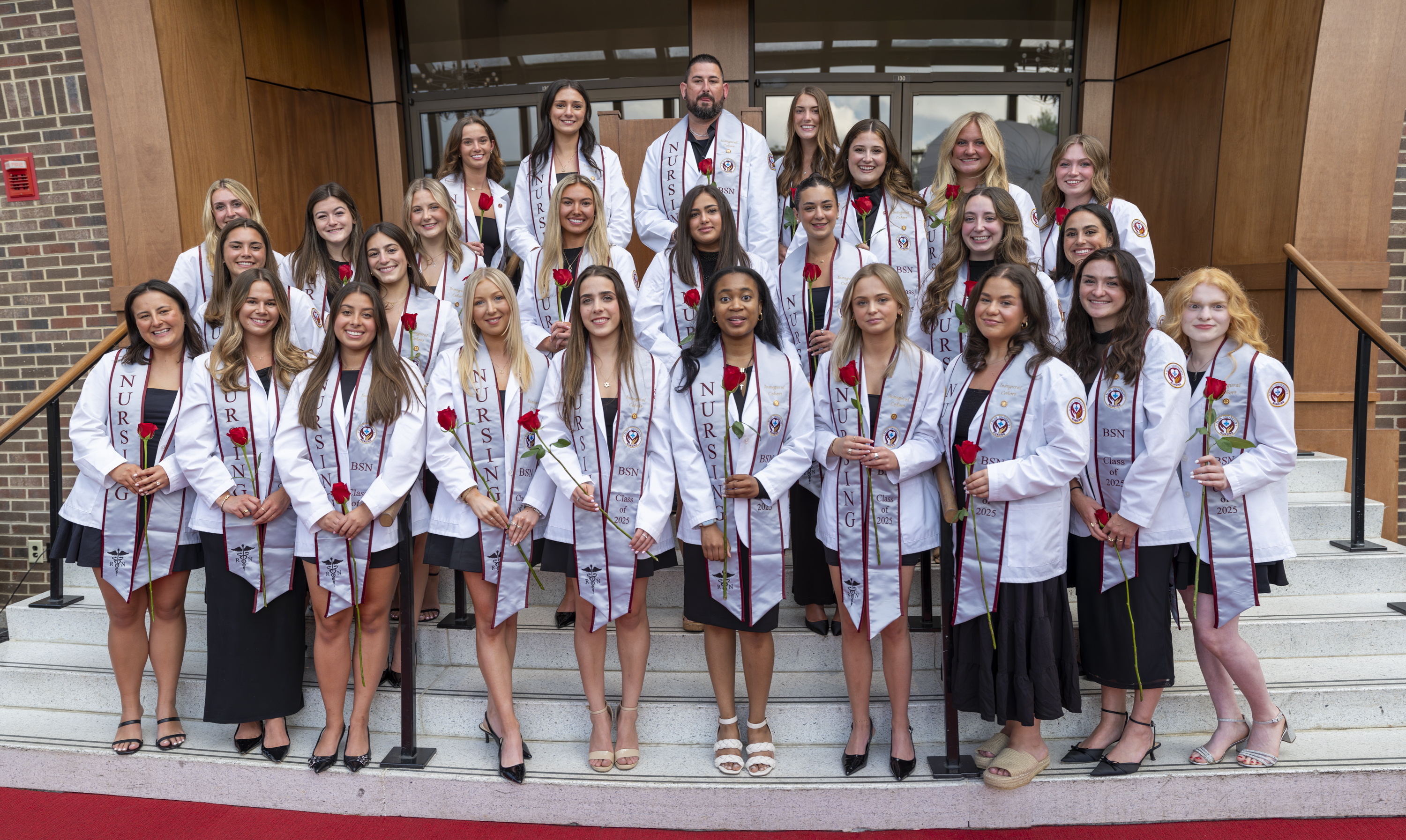 The inaugural cohort of Elon’s Bachelor of Science in Nursing program poses in white coats and stoles on the steps outside a campus building.