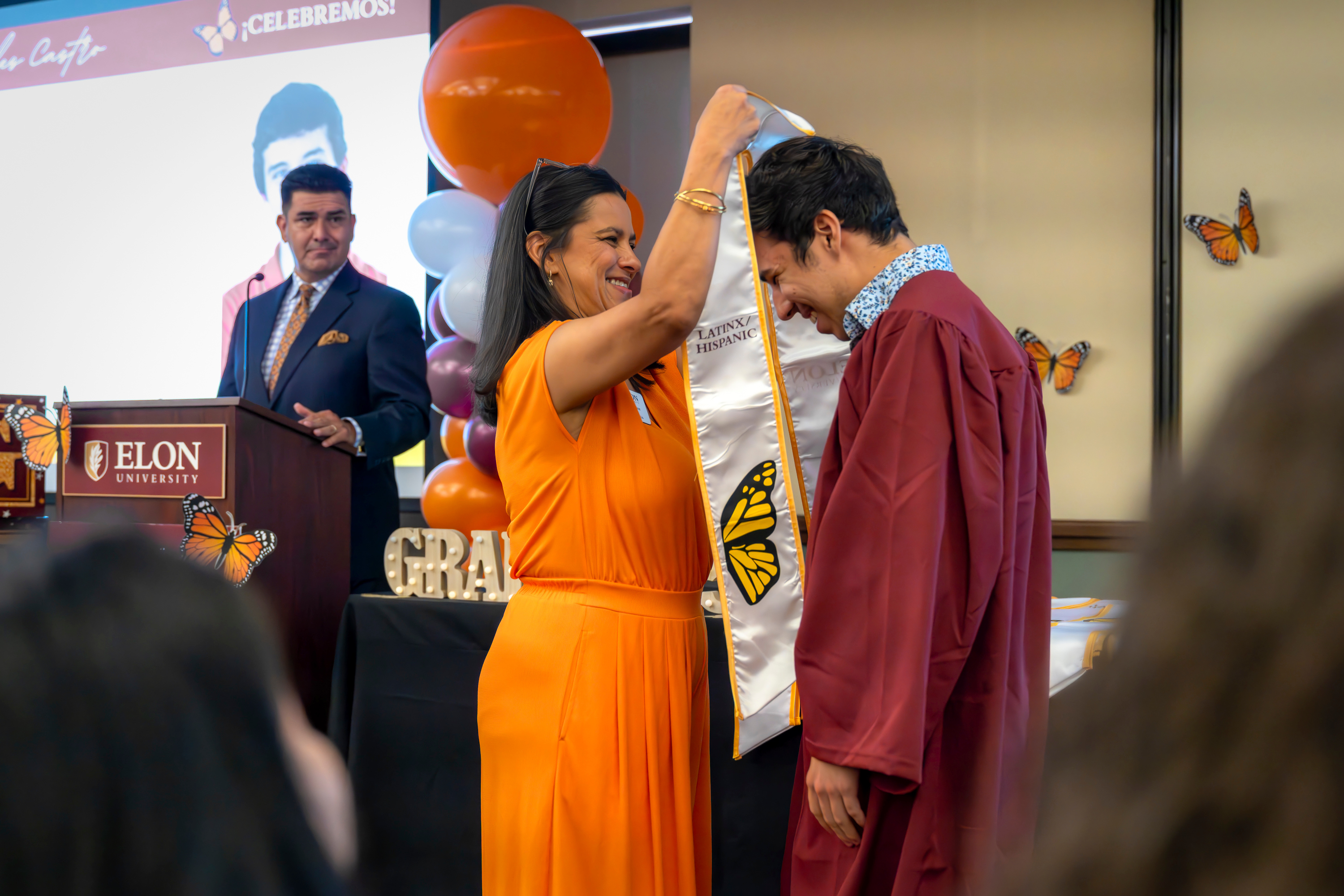 Assistant Dean of Students and CREDE Director Sylvia Muñoz, in an orange dress, places a white stole with a monarch butterfly design over the shoulders of a smiling graduate in a maroon cap and gown, Charles Castro, during Elon University’s ¡Celebremos! ceremony. A man stands at a podium in the background with an Elon University sign, and festive balloons and butterfly decorations surround the stage.