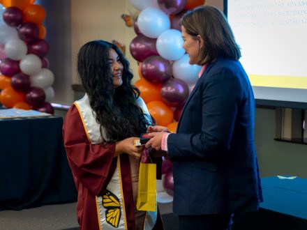 Rachel Lopez, an Elon graduate in a maroon cap and gown with a white stole featuring a monarch butterfly, smiles while receiving a small gift bag from President Connie Ledoux Book during Elon University’s ¡Celebremos! ceremony.