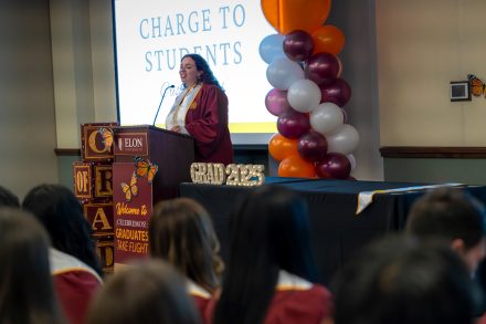Wearning a maroon cap and gown, Cristy Mariné stands at a podium delivering the "Charge to Students" during Elon University’s ¡Celebremos!: Graduates Take Flight ceremony.
