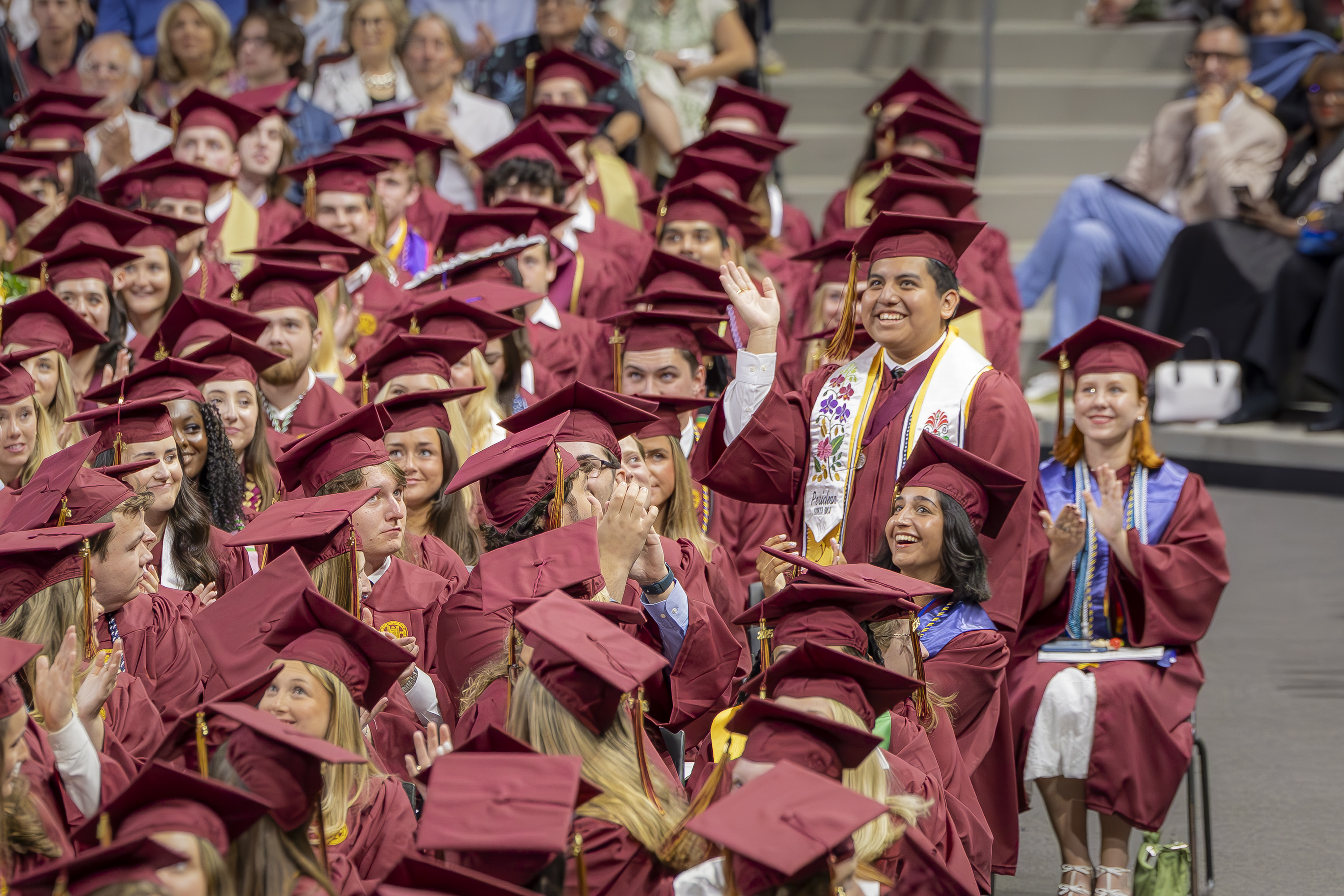 A graduate in maroon regalia stands and waves with a joyful smile amid a crowd of seated classmates, as applause and cheers erupt around him during the ceremony.
