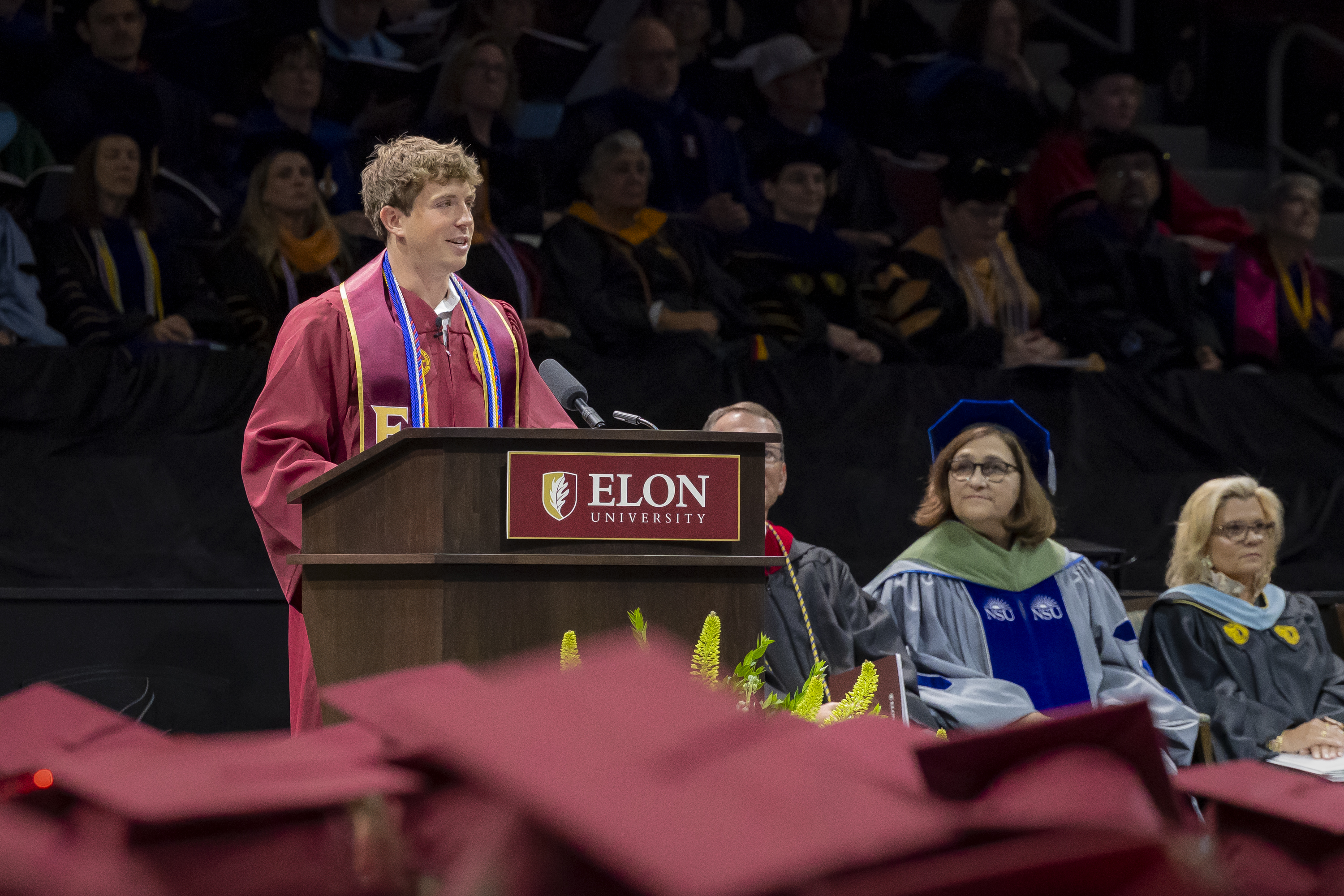 A student speaker in graduation regalia addresses the audience from a podium at Elon University’s Commencement ceremony.