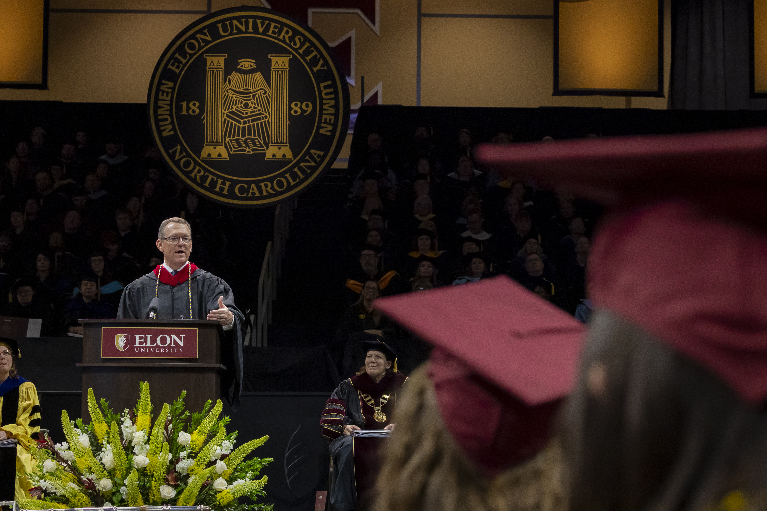 A speaker gestures while delivering remarks onstage at Commencement, with the Elon University seal displayed prominently behind them.