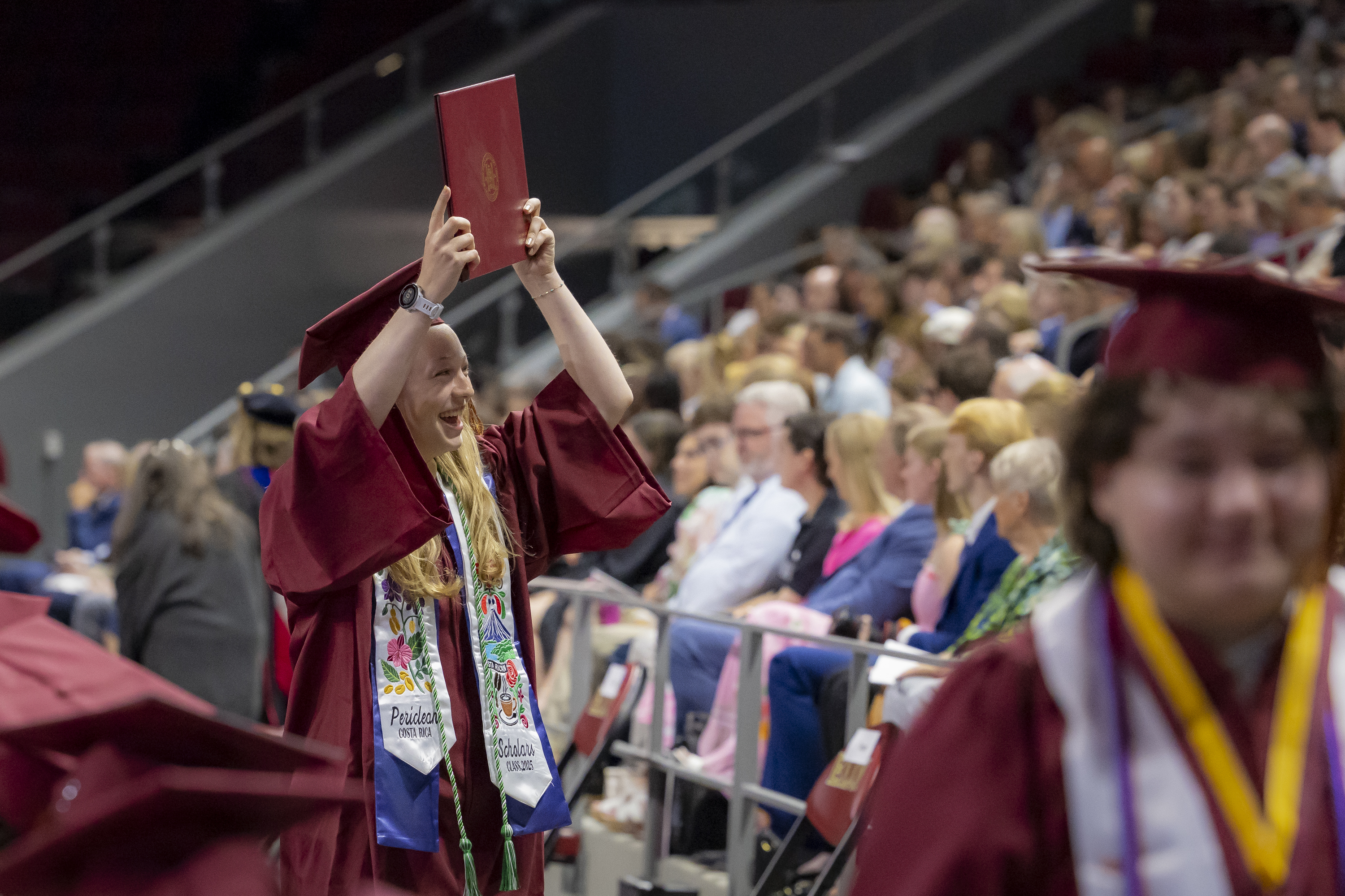 A jubilant graduate raises her diploma high with a beaming smile as she walks through the crowd.