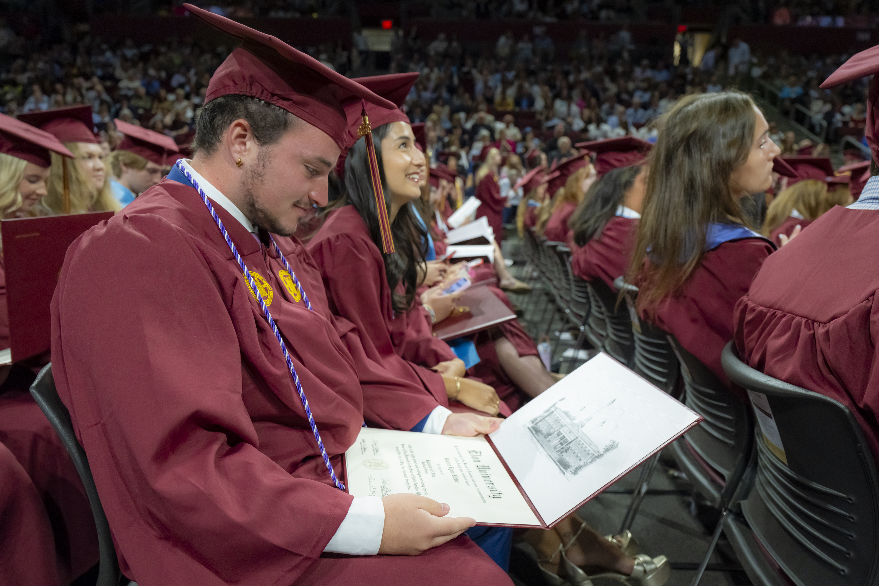 Graduates seated in rows smile and admire their diplomas during Elon University’s Commencement ceremony, surrounded by fellow classmates and an audience of proud supporters.