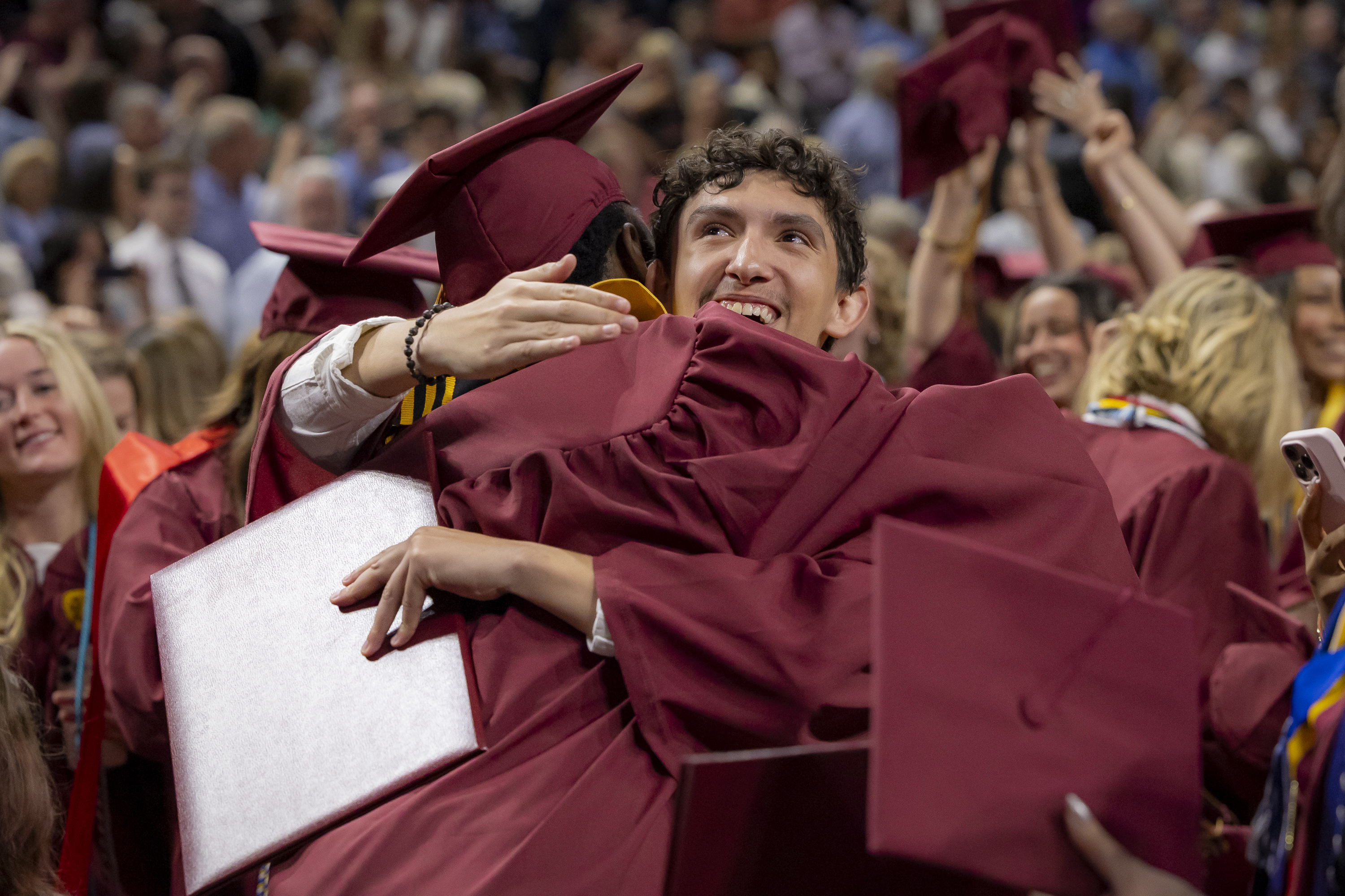 6. A graduate in maroon regalia waves to the audience while crossing the stage during the Commencement ceremony. 7. Elon University President Connie Ledoux Book smiles widely while holding up a symbolic oak sapling gift during her Commencement address. 8. Two graduates embrace joyfully in a sea of maroon caps and gowns during the celebration.