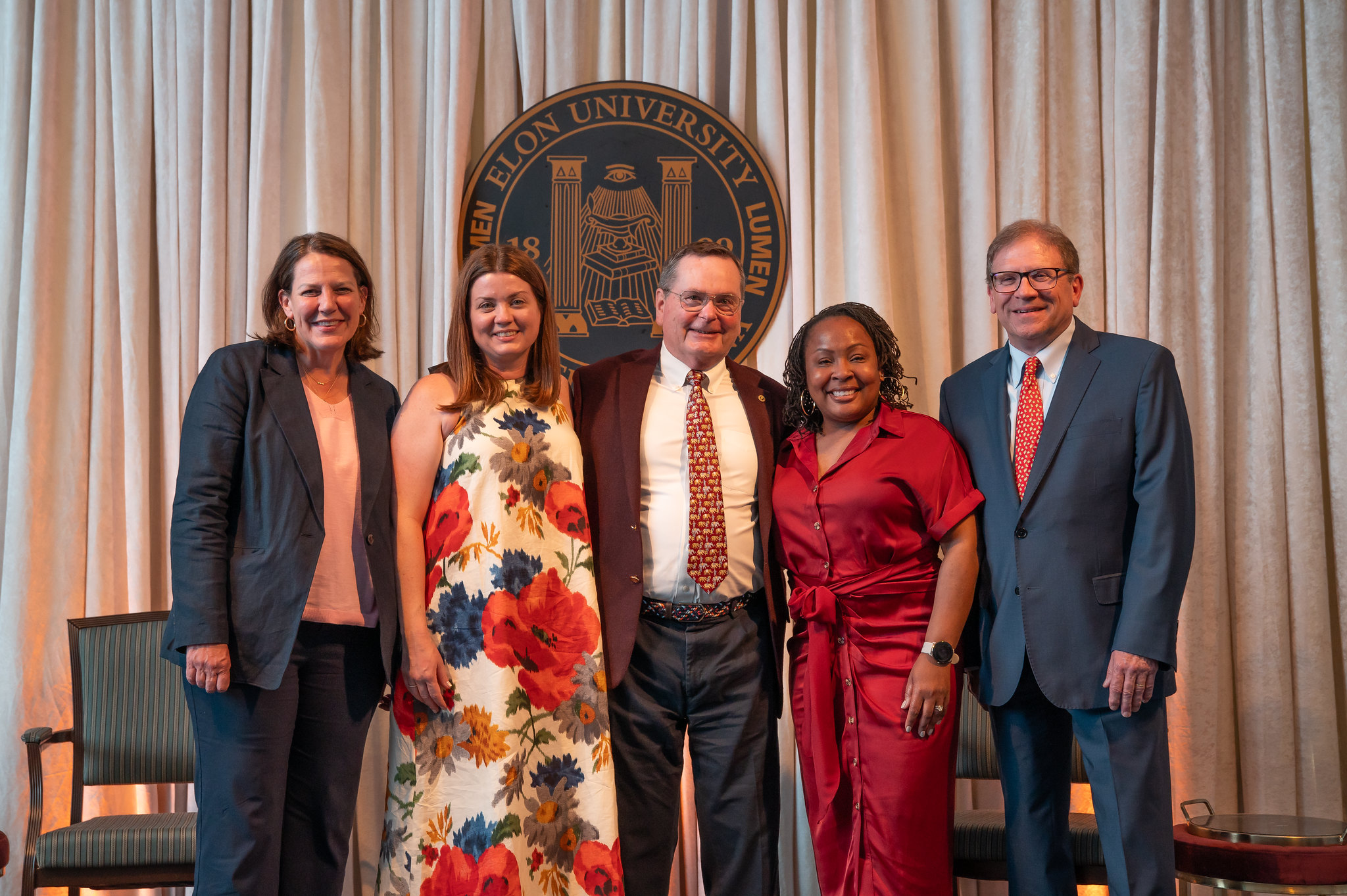 President Connie Ledoux Book stands with the 2025 Distinguished Alumni Awards recipients—Cecelia Thompson, Sam Troy, Akilah Weaver Hill, and Mike Cross—posing in front of an Elon University seal on stage with beige curtains in the background. All are smiling and dressed in professional or semi-formal attire.