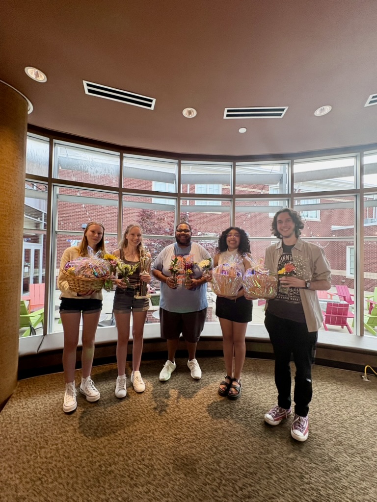 Five smiling people stand in front of large windows holding colorful gift baskets, with a brick courtyard visible outside.