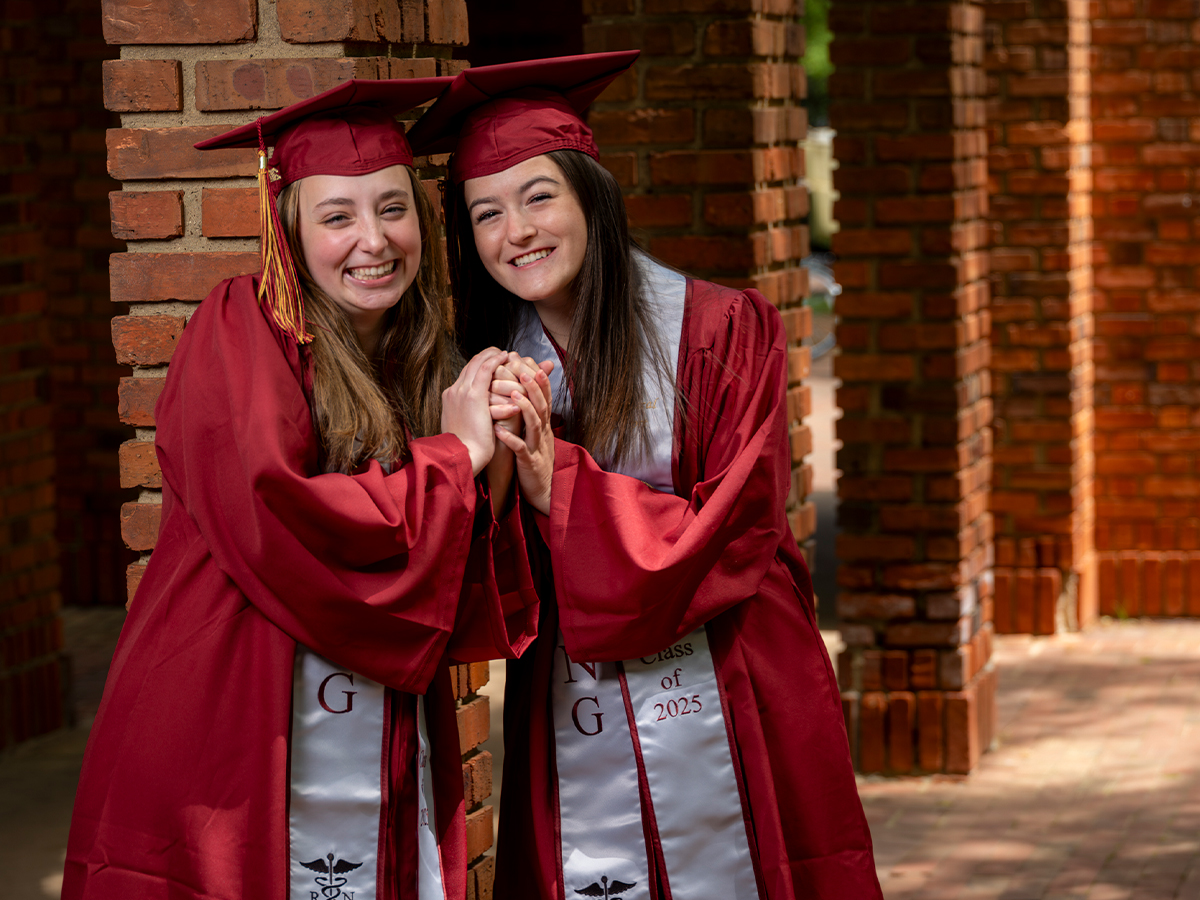 Two nursing graduates in matching maroon caps and gowns clasp hands and smile joyfully in front of brick columns.