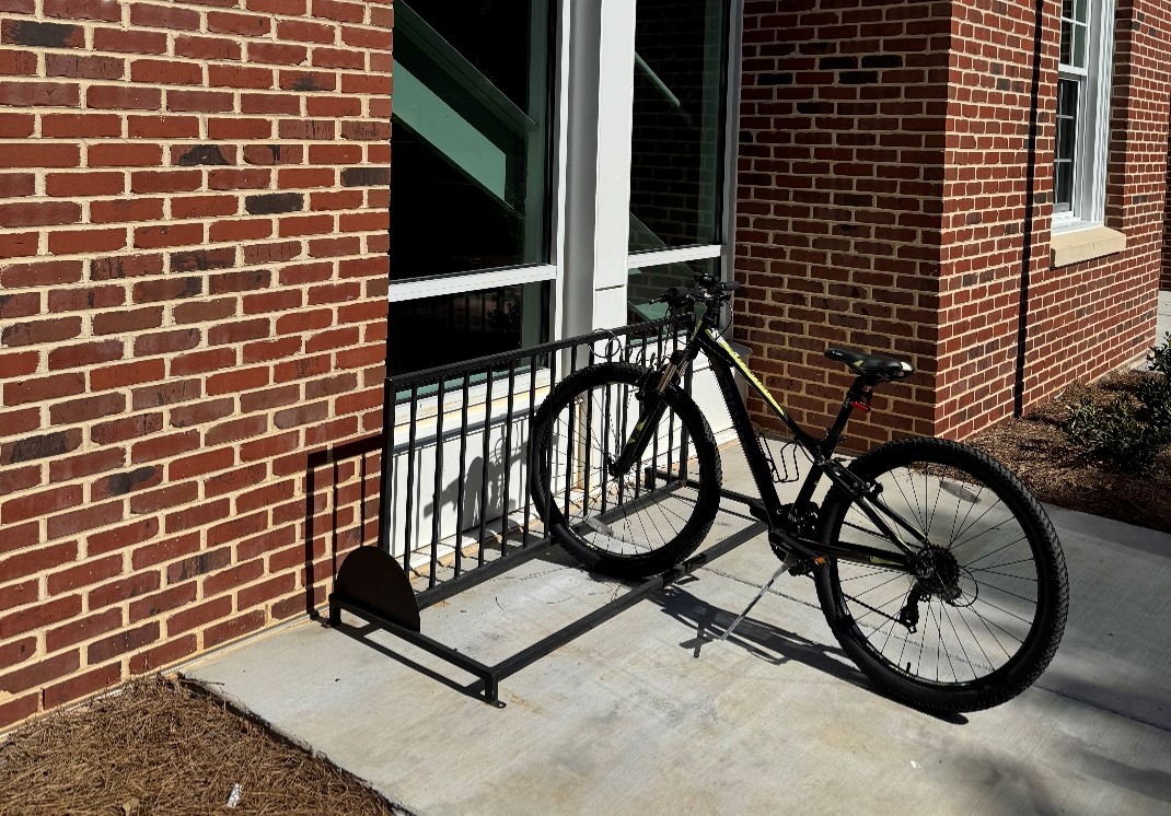 A black bicycle is parked in a metal bike rack outside a red-brick building near a large window.