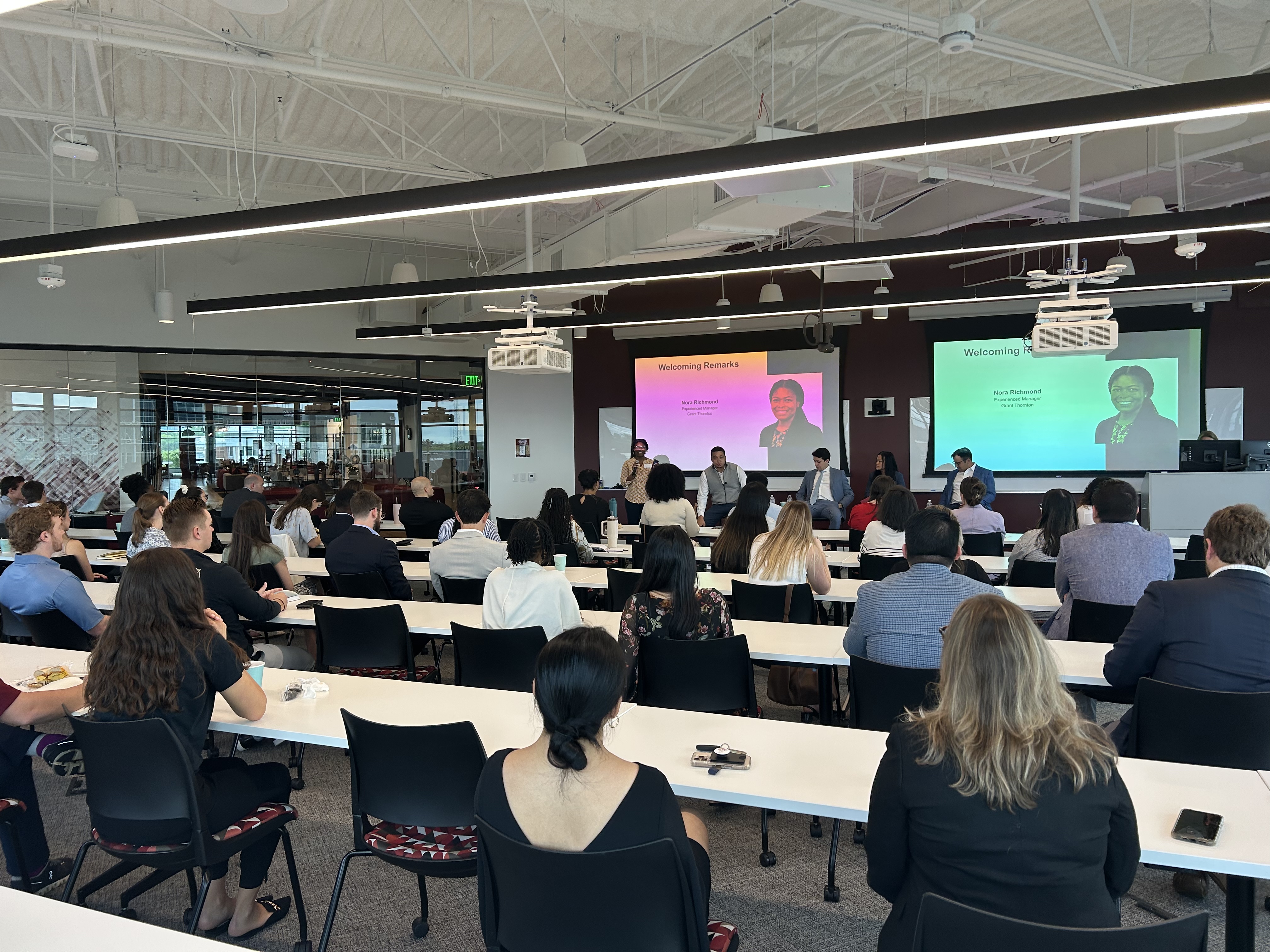 Attendees seated in a modern classroom listen to a panel discussion, with a presentation projected on two screens at the front of the room