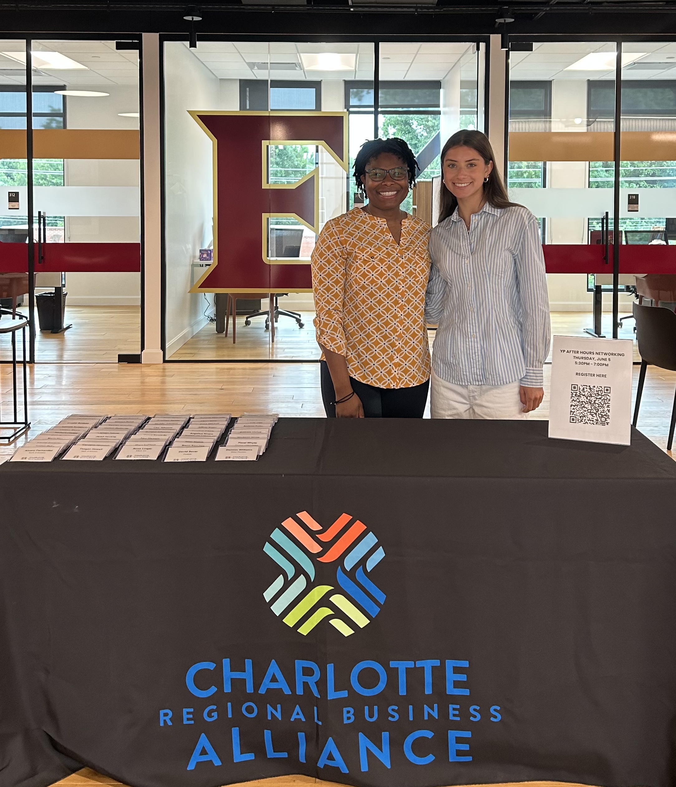 Two women stand behind a Charlotte Regional Business Alliance table at a networking event, with a large maroon and gold ‘E’ visible through the glass wall behind them.