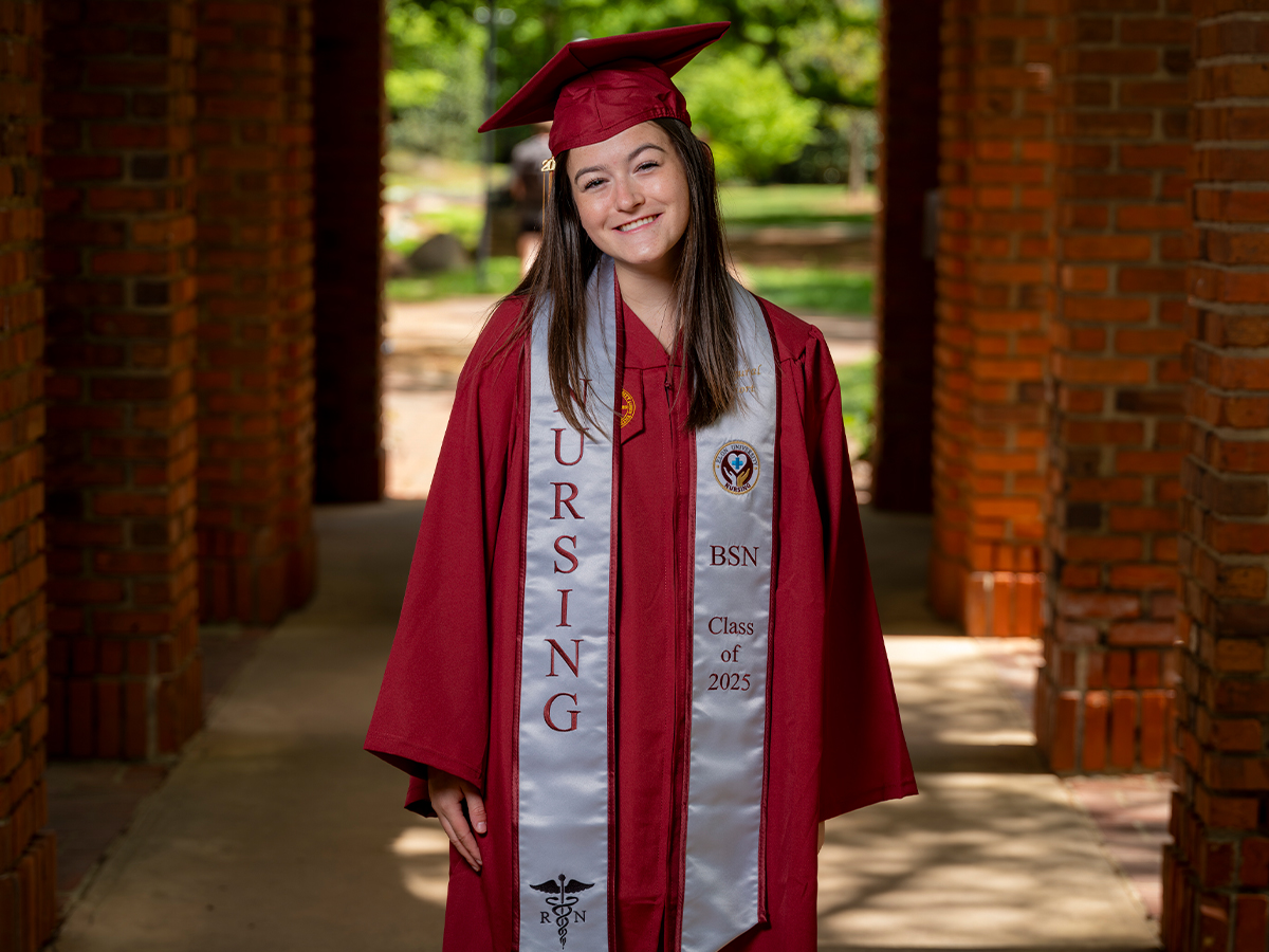 A nursing graduate in a maroon cap and gown stands proudly in a brick corridor with a stole that reads “NURSING BSN Class of 2025.”
