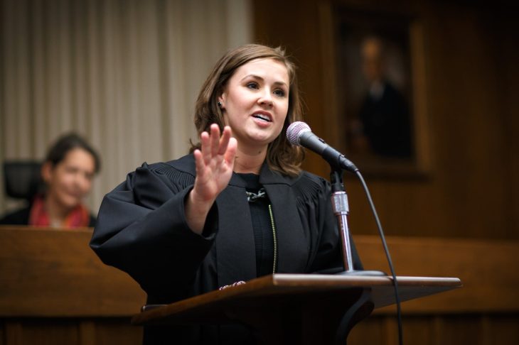 A woman in judicial robes speaks passionately at a podium with a microphone in a courtroom. A blurred figure is seated in the background, and a framed portrait hangs on the wood-paneled wall behind her.