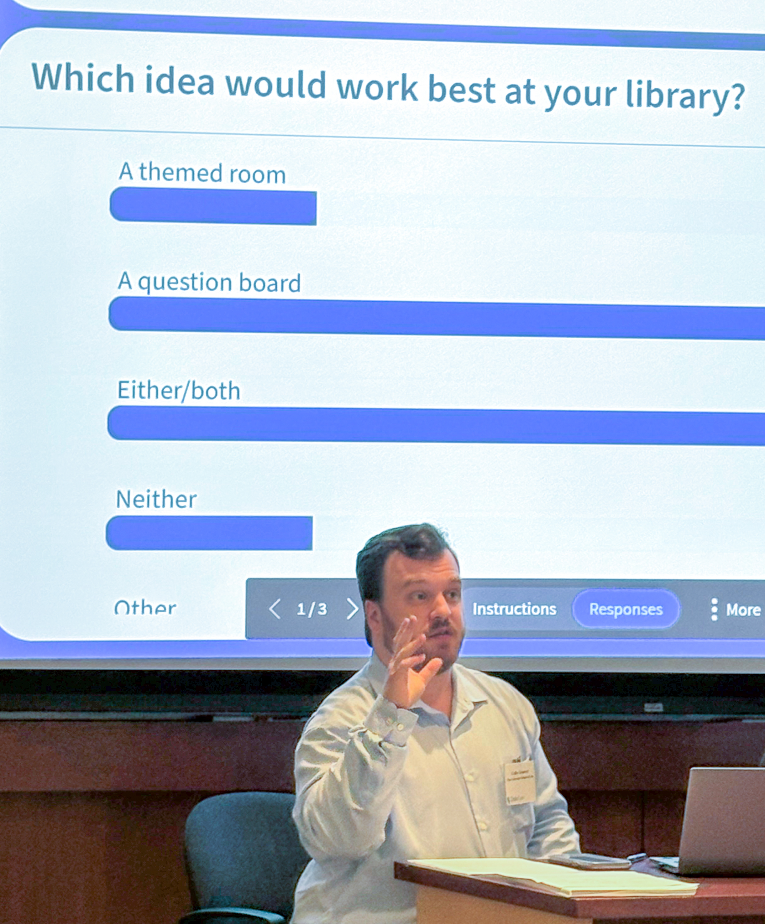 A man seated at a desk gestures while speaking. A chart is shown on a projected screen behind him.