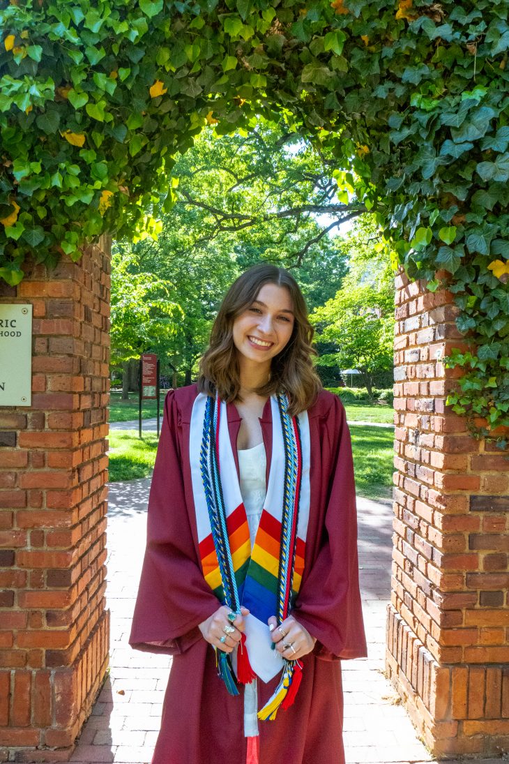 This is a celebratory graduation photo. The image features a young woman standing outdoors under a brick archway covered in green ivy. She is smiling and dressed in a maroon graduation gown with a white dress underneath. Around her neck are multiple honor cords and stoles, including a prominent stole with a rainbow design, which often represents LGBTQ+ pride. The setting appears to be on a college or university campus, with lush green trees and brick pathways in the background, suggesting a warm, sunny day.
