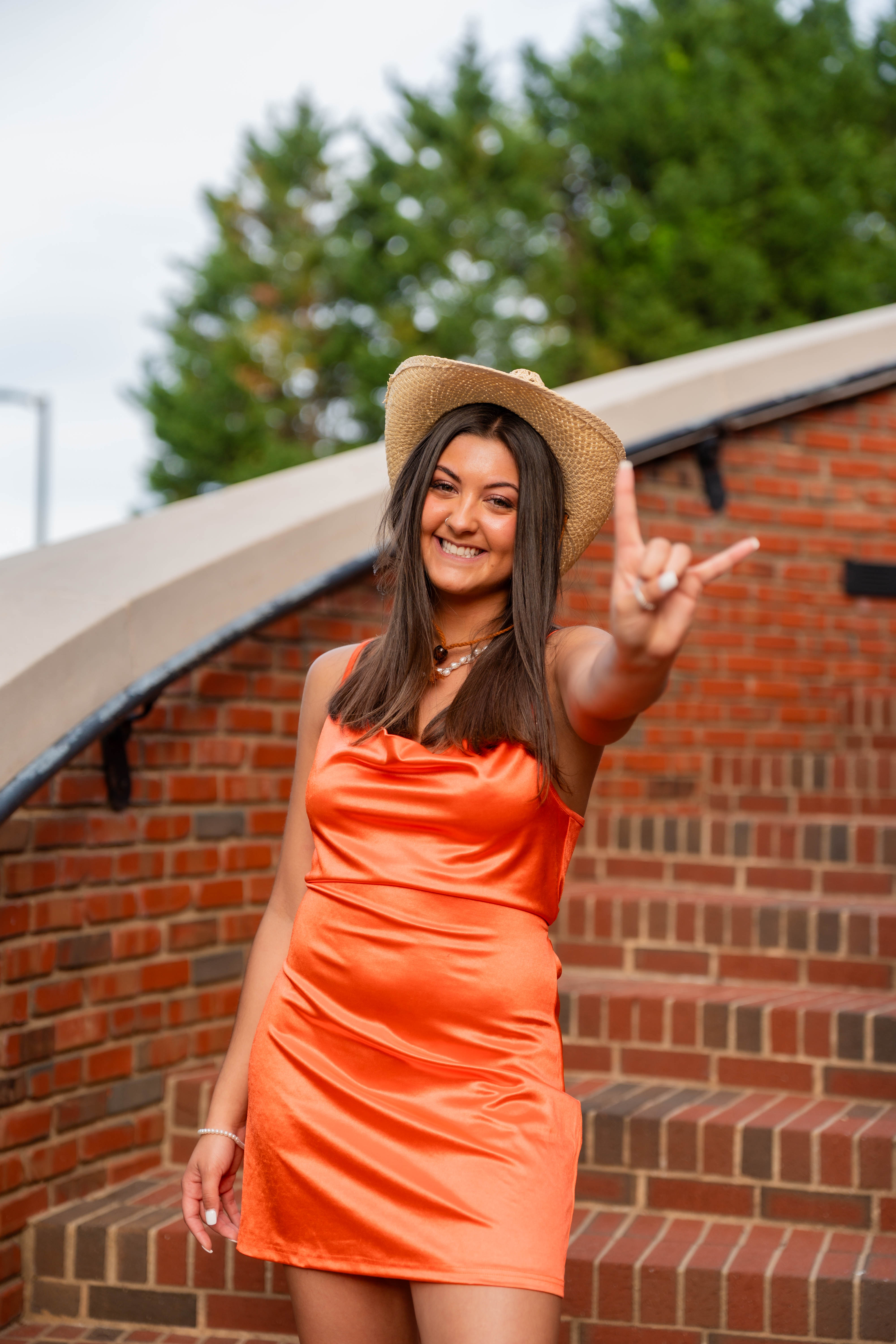 A young woman in an orange dress and cowboy hat smiles while making a hand gesture, standing confidently on a brick staircase with greenery in the background.