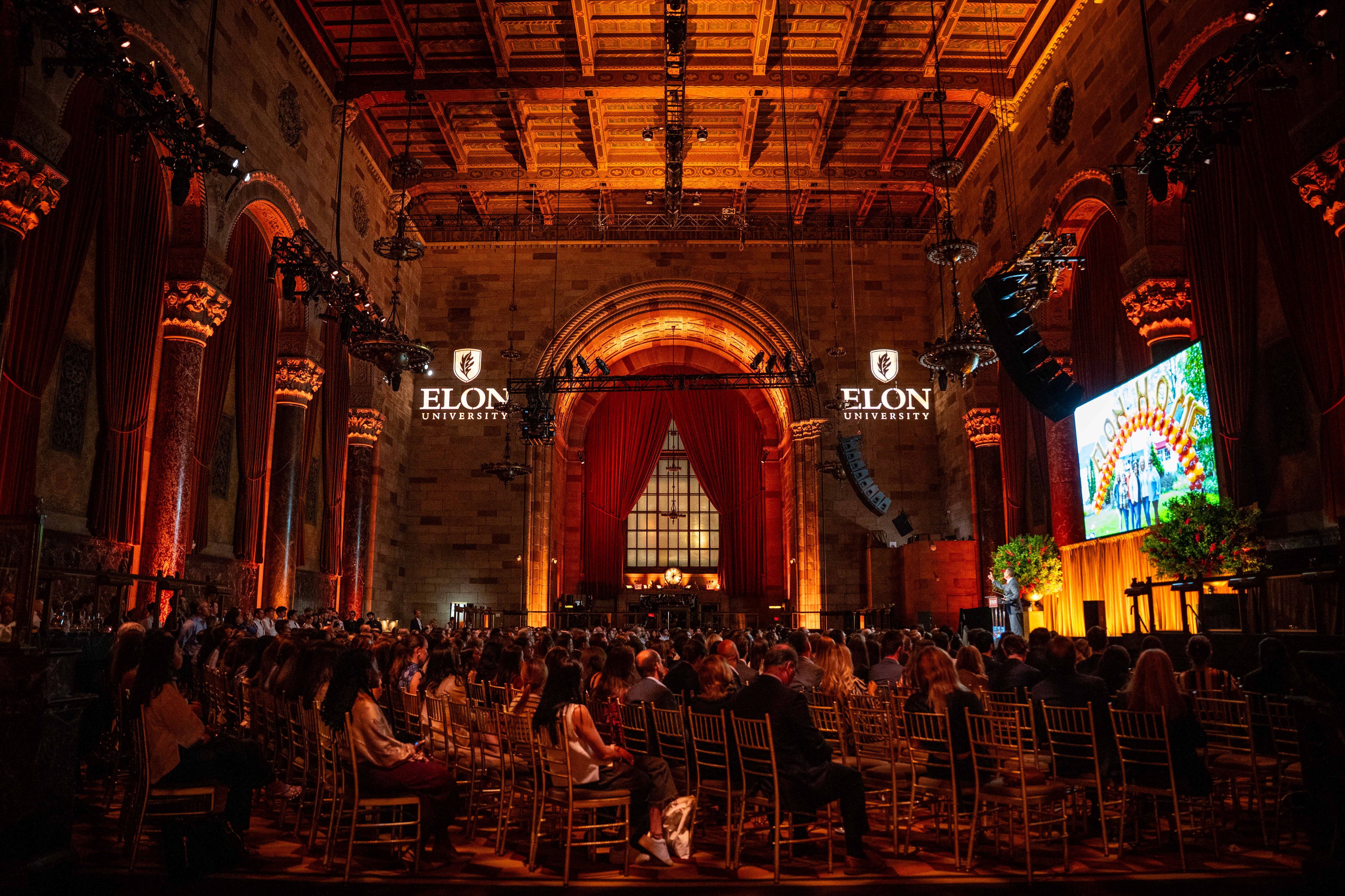 A warmly lit, ornate auditorium displays two illuminated “Elon University” logos on the stone walls, with a large screen showing a colorful campus image on the right