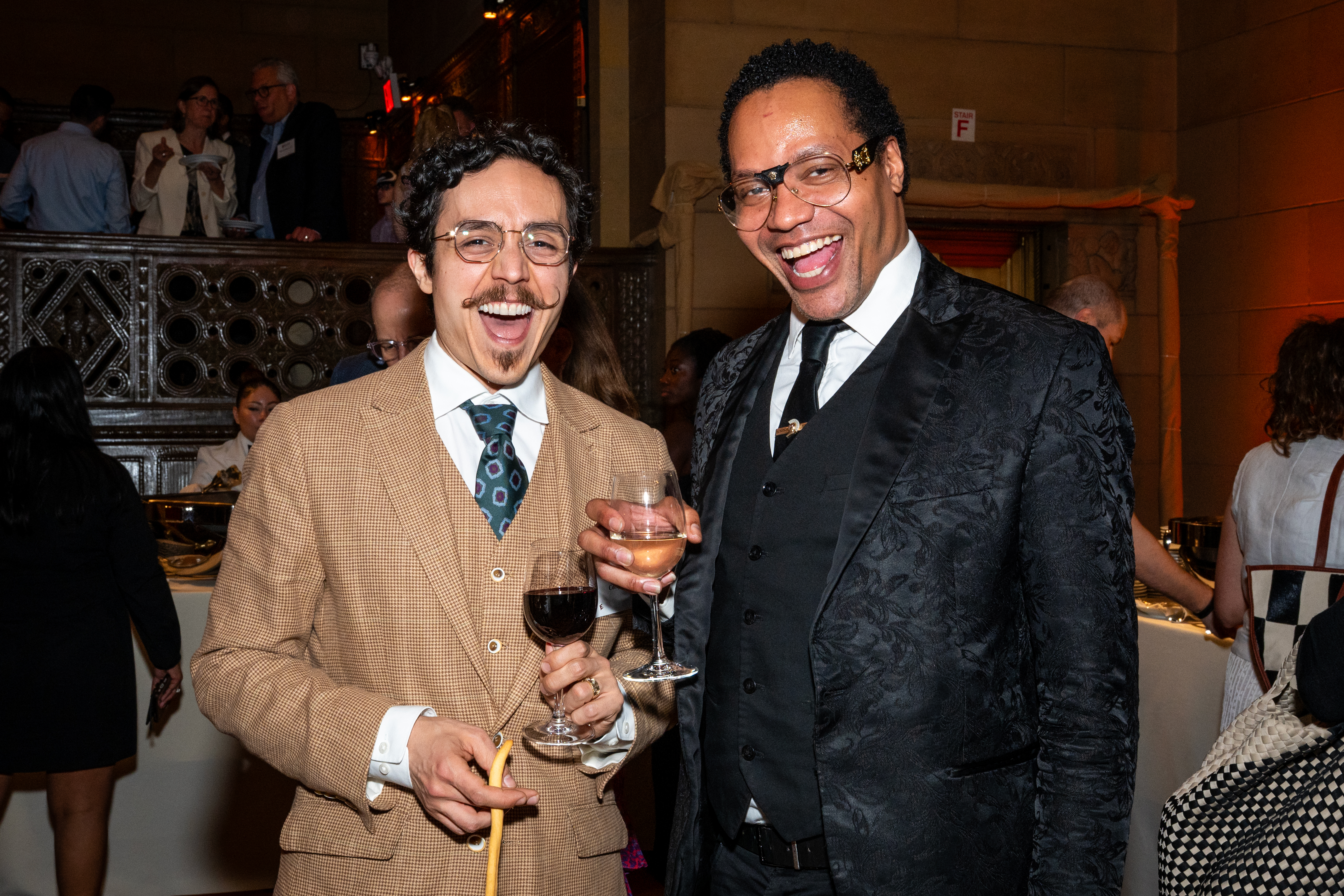Two well-dressed men smile and raise glasses of wine at a formal event, surrounded by other attendees in an elegant venue.