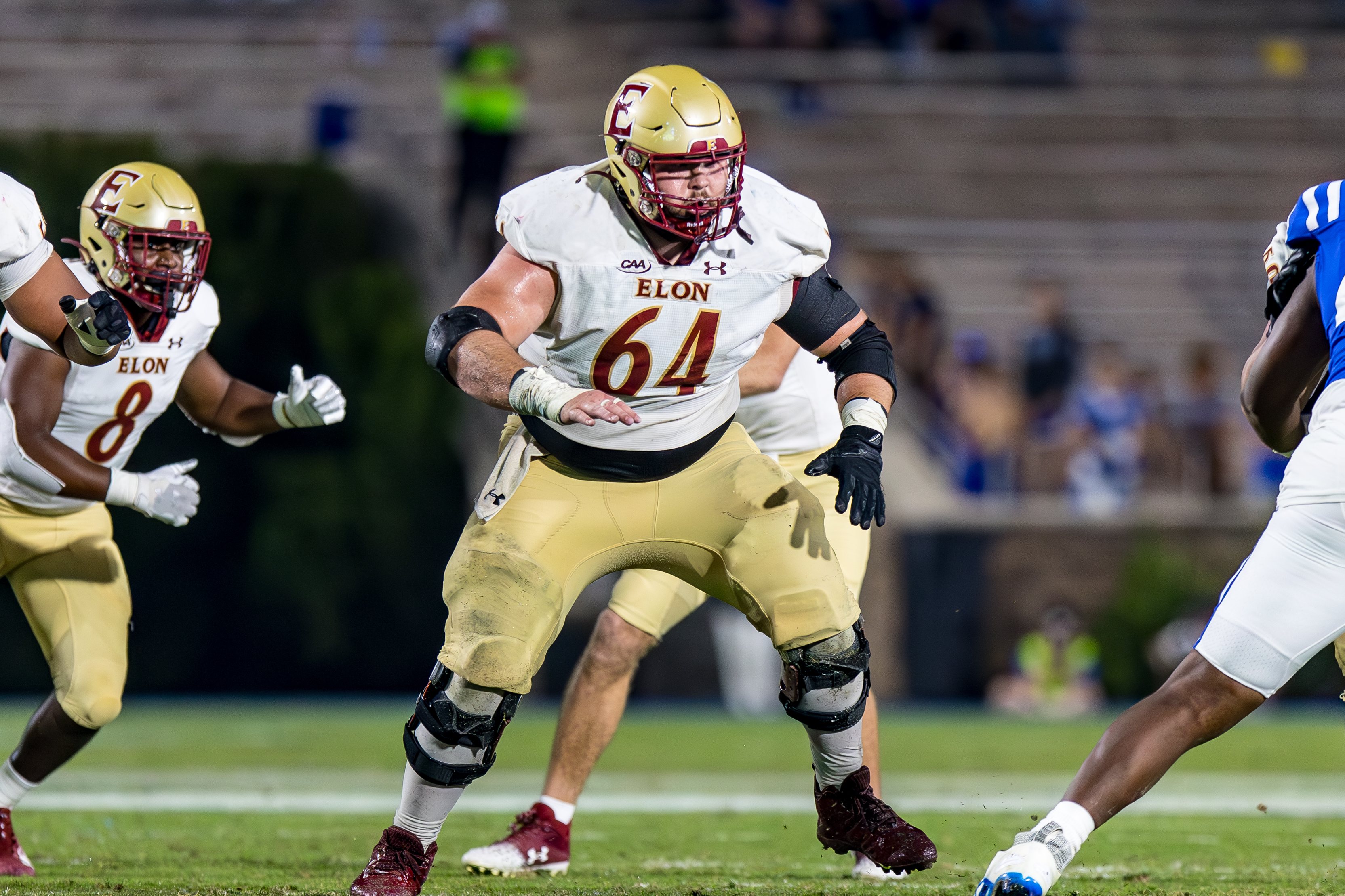 An Elon University football player wearing jersey number 64 prepares to block an opponent during a game, with teammates and players from the opposing team visible in the background.
