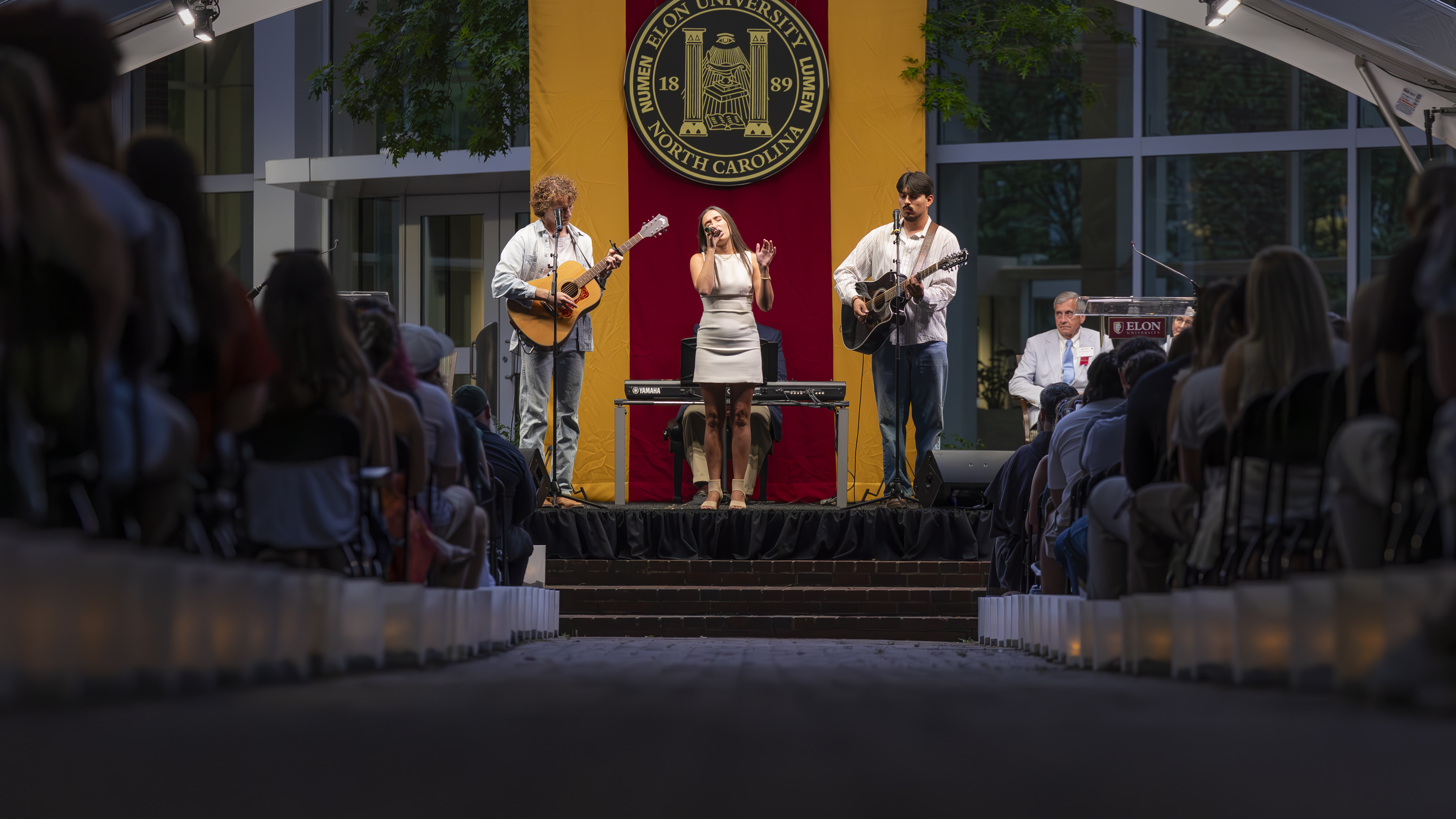 Three student musicians perform a song on stage during Elon’s Numen Lumen ceremony, framed by the university seal and surrounded by rows of candlelit attendees.