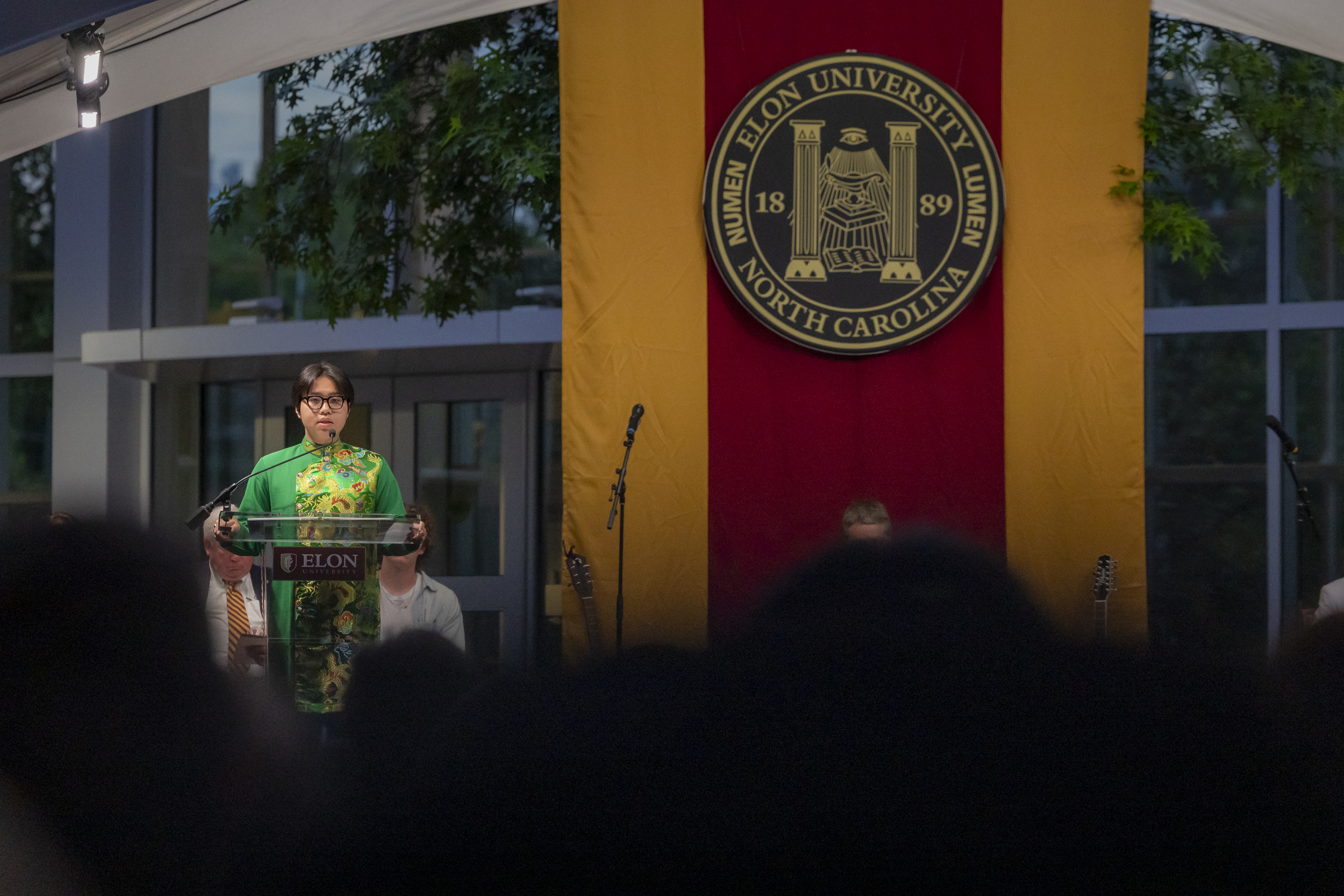 A student speaker stands at the podium in traditional attire during Elon University’s Numen Lumen ceremony, with the university seal displayed prominently behind them.