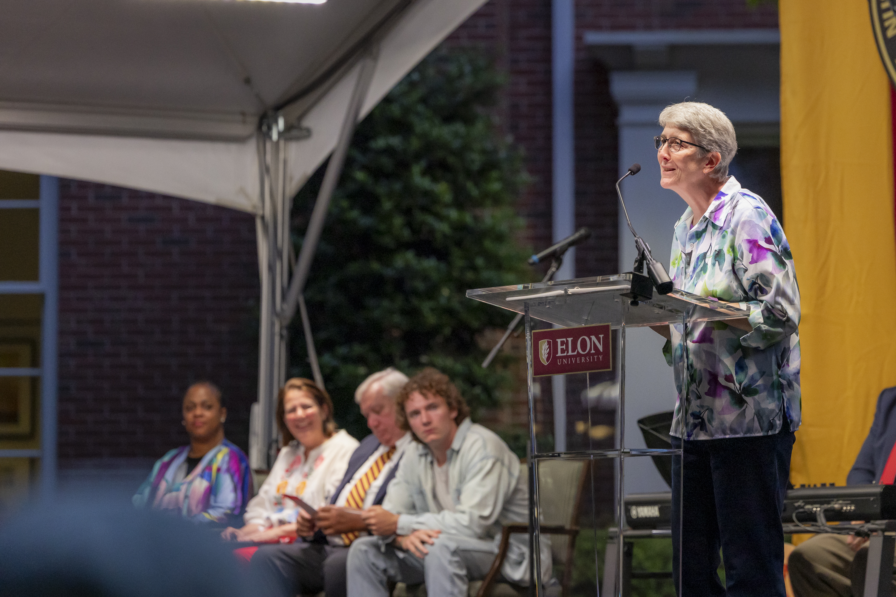 Professor Mary Jo Festle speaks at the podium during Elon’s Numen Lumen Senior Baccalaureate Reflection, with fellow faculty and guests seated behind her on stage.
