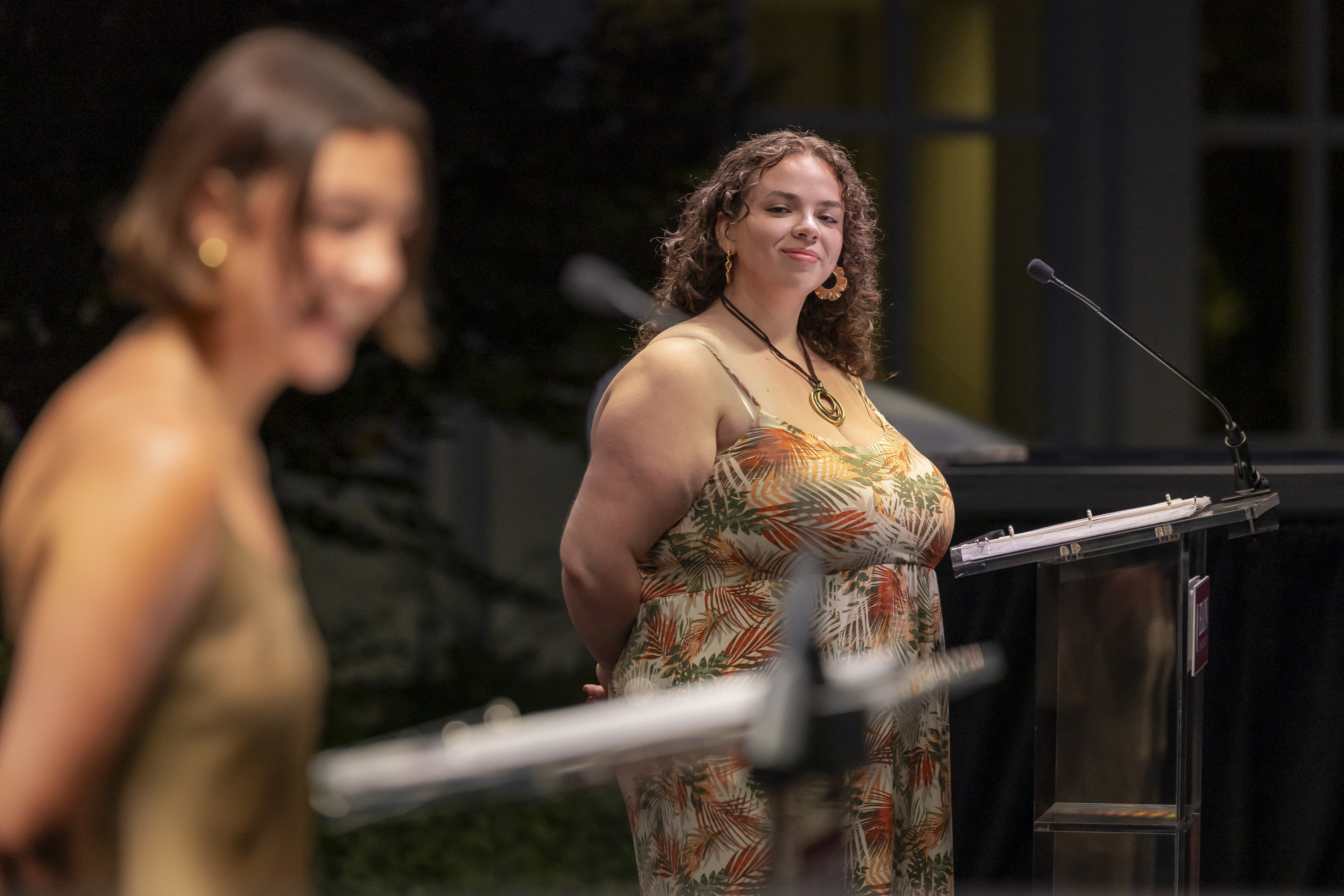 A student stands confidently at the podium during Elon’s Numen Lumen ceremony, smiling as another speaker presents in the foreground.