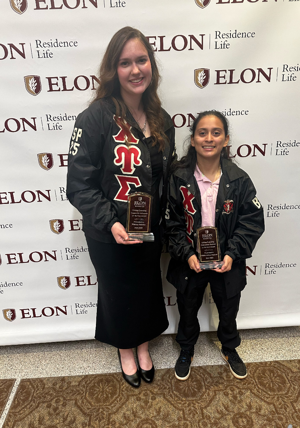 Two women wearing matching Greek life jackets hold awards and smile in front of an Elon University Residence Life step-and-repeat banner.