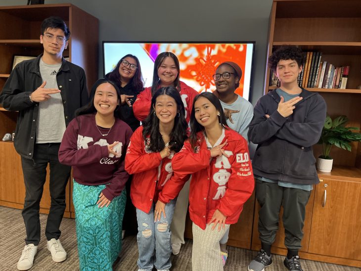 This photo features a group of eight young adults posing together indoors, likely in a student center or similar setting. They are smiling and making various hand signs, suggesting a sense of camaraderie, friendship, or affiliation with a club or organization. Key details: The background includes wooden shelves with books, a potted plant, and a television screen displaying red and white imagery, possibly festive or cultural in theme. Three of the people in the front row (center and right) are wearing matching red jackets with Greek letters, indicating that they might be members of a fraternity or sorority, specifically Kappa Phi Lambda (ΚΦΛ), a historically Asian-interest sorority. Another person in the front row wears a maroon sweatshirt also featuring Greek letters, possibly representing a different organization. The group's overall demeanor is cheerful and relaxed, suggesting they are celebrating or commemorating an event together.